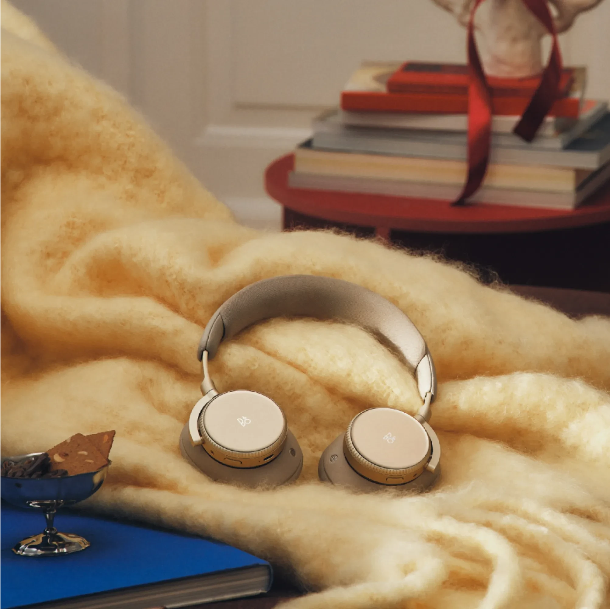 Beoplay H100 sand wireless headphones resting on a soft cream blanket with stacked books and a red ribbon in the background.