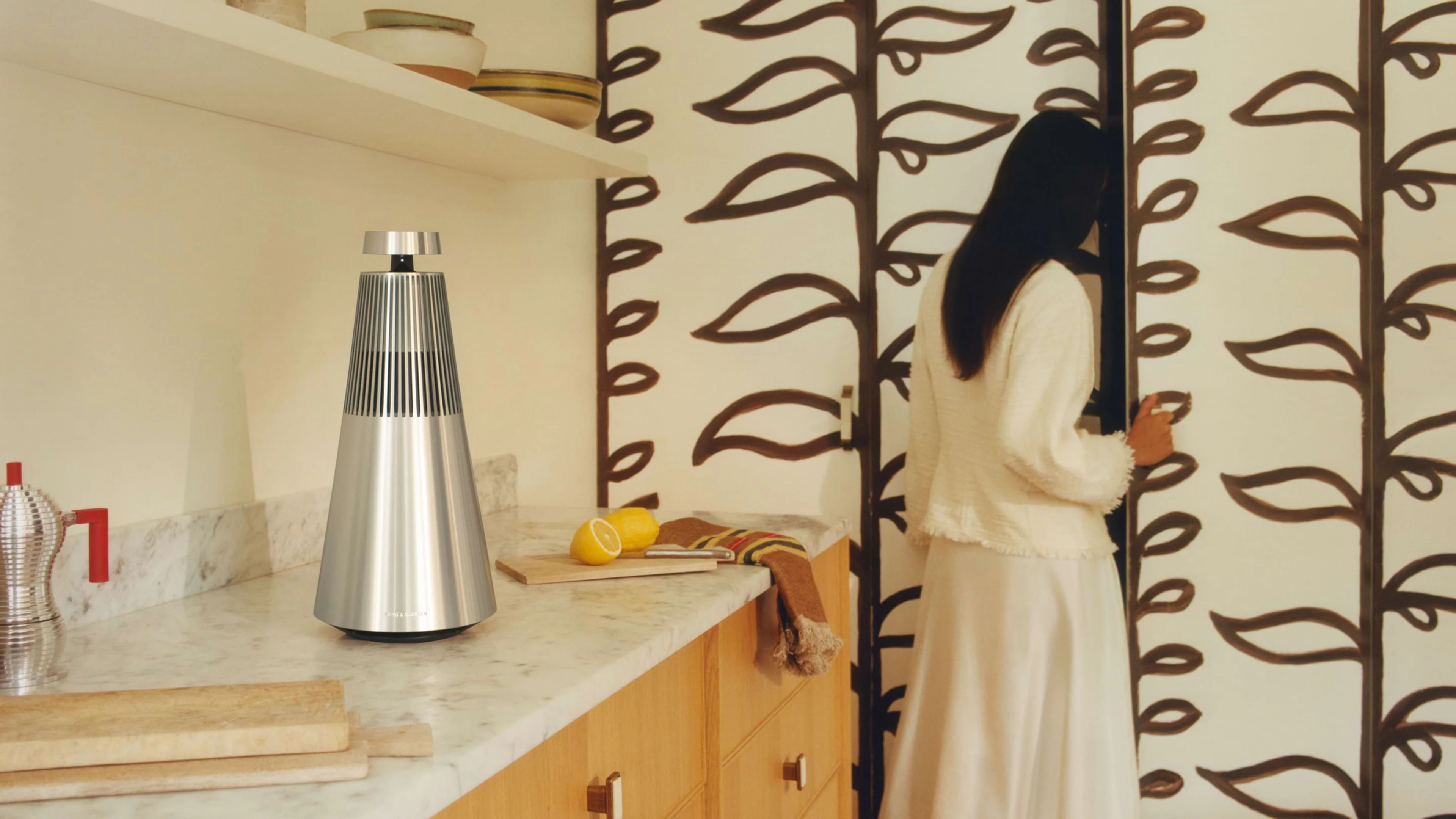 Beosound 2 on a marble furniture in the kitchen and the lady is looking for something inside the cabinet