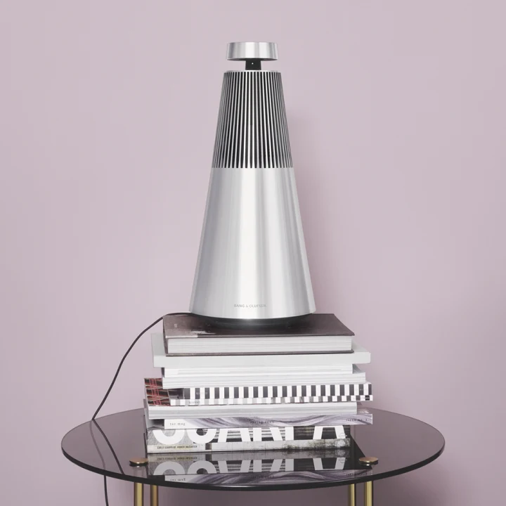 Beosound 2 in natural aluminium on a stack of magazines in front of a lilac background wall
