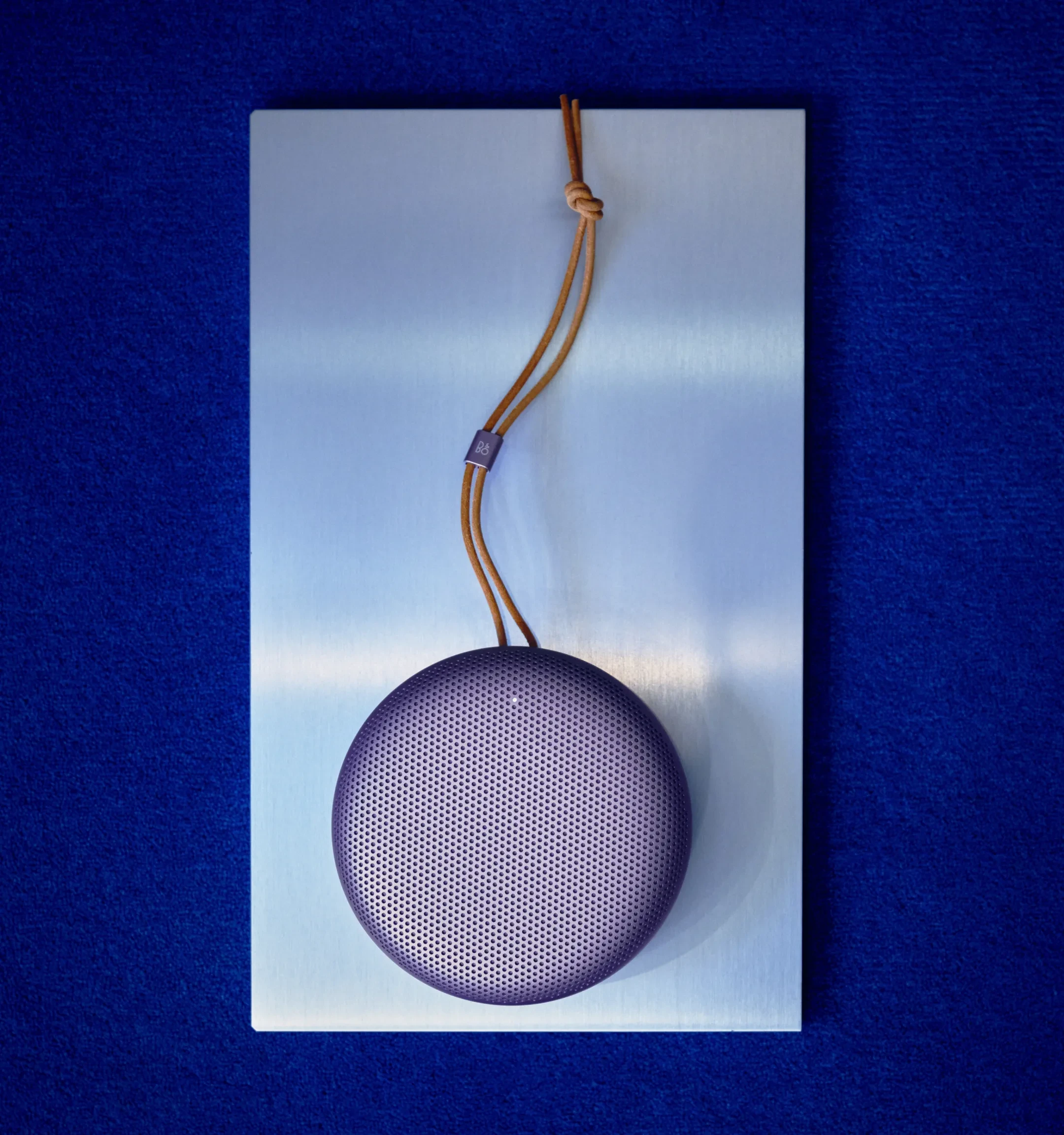 Beosound A1, 3rd generation, in the color variant Lavender Purple, laying on the metal table with blue background