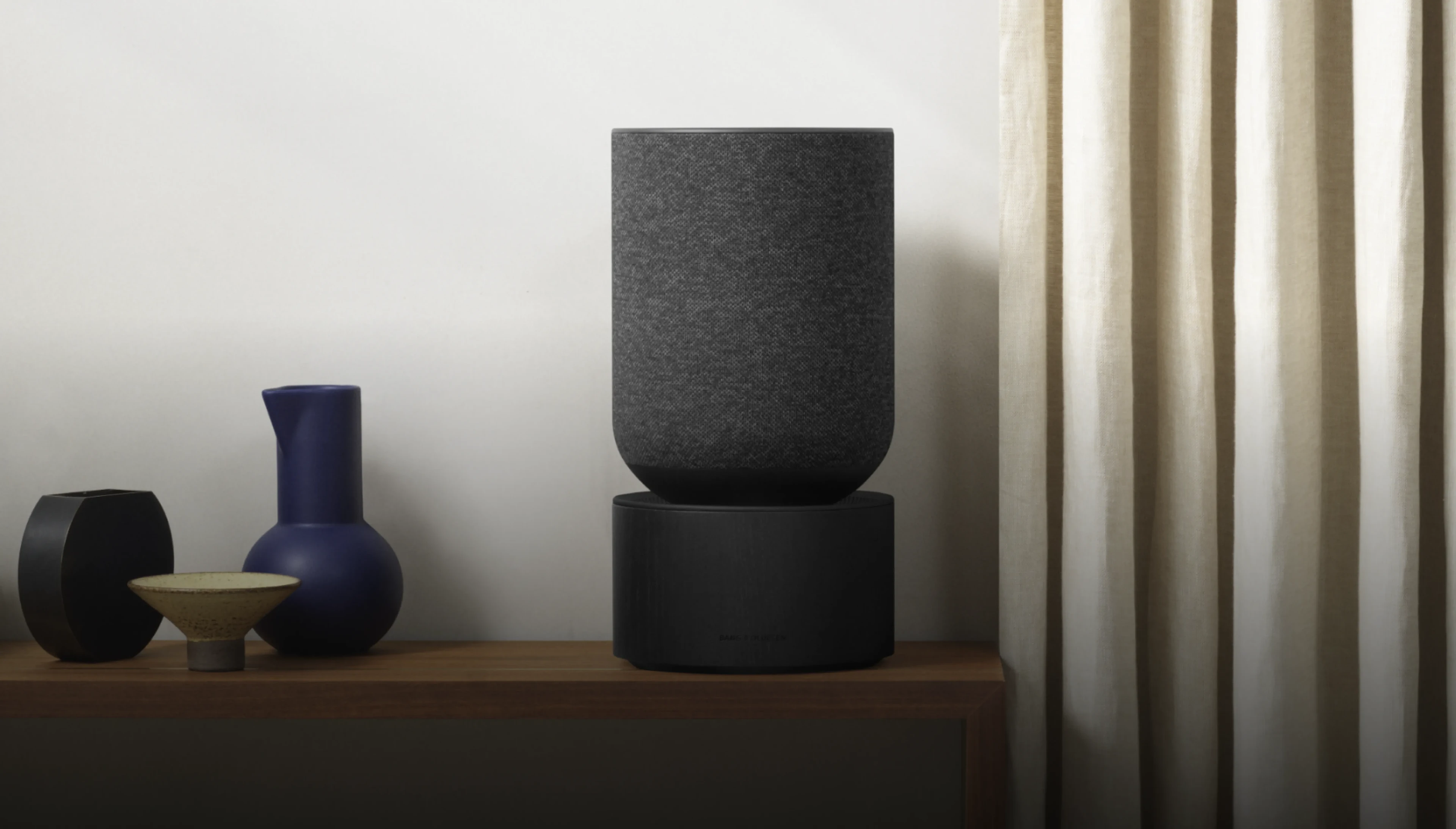 Beosound Balance displayed on a wooden shelf in a room with curtains and vase.