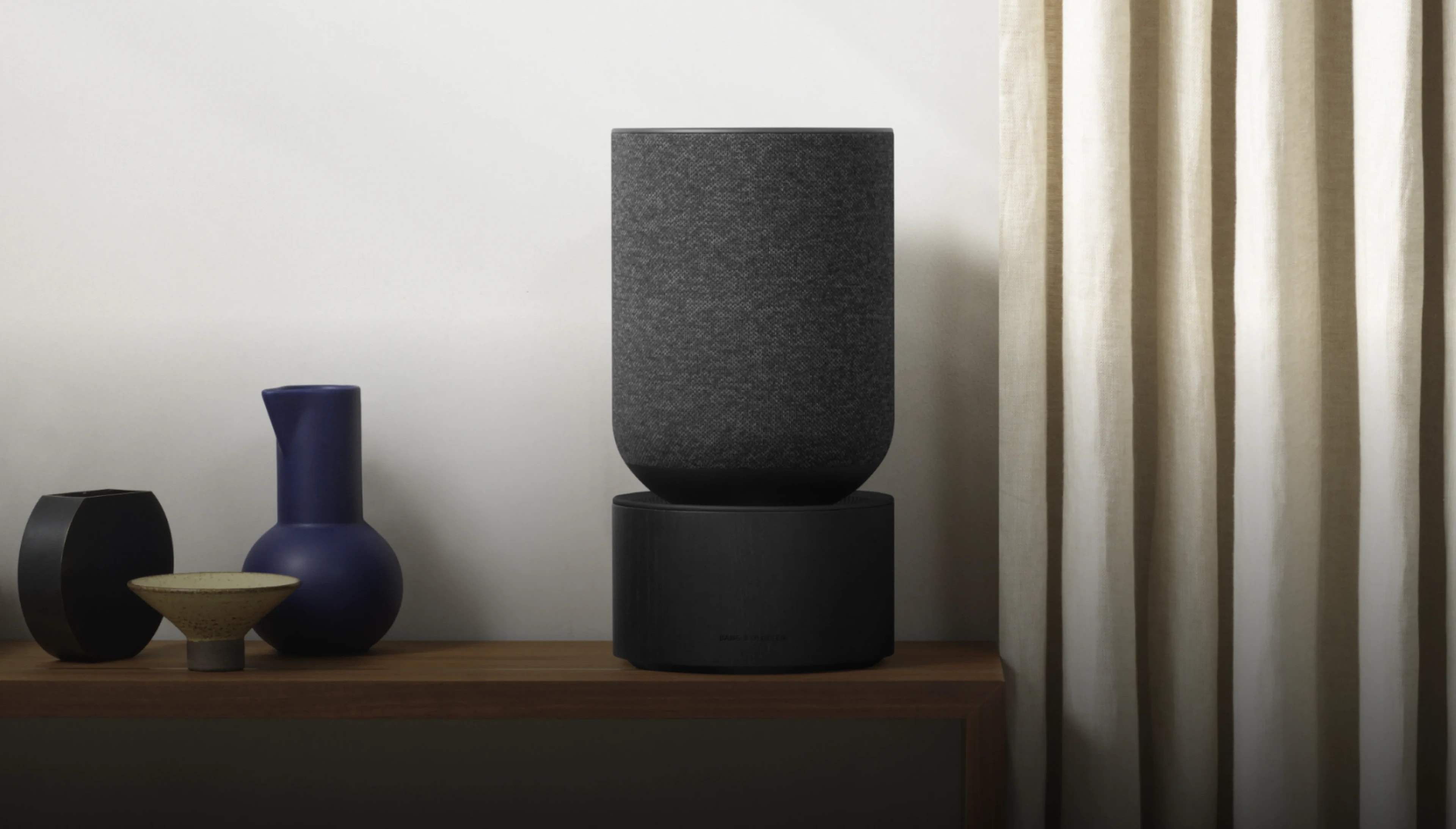 Beosound Balance displayed on a wooden shelf in a room with curtains and vase.