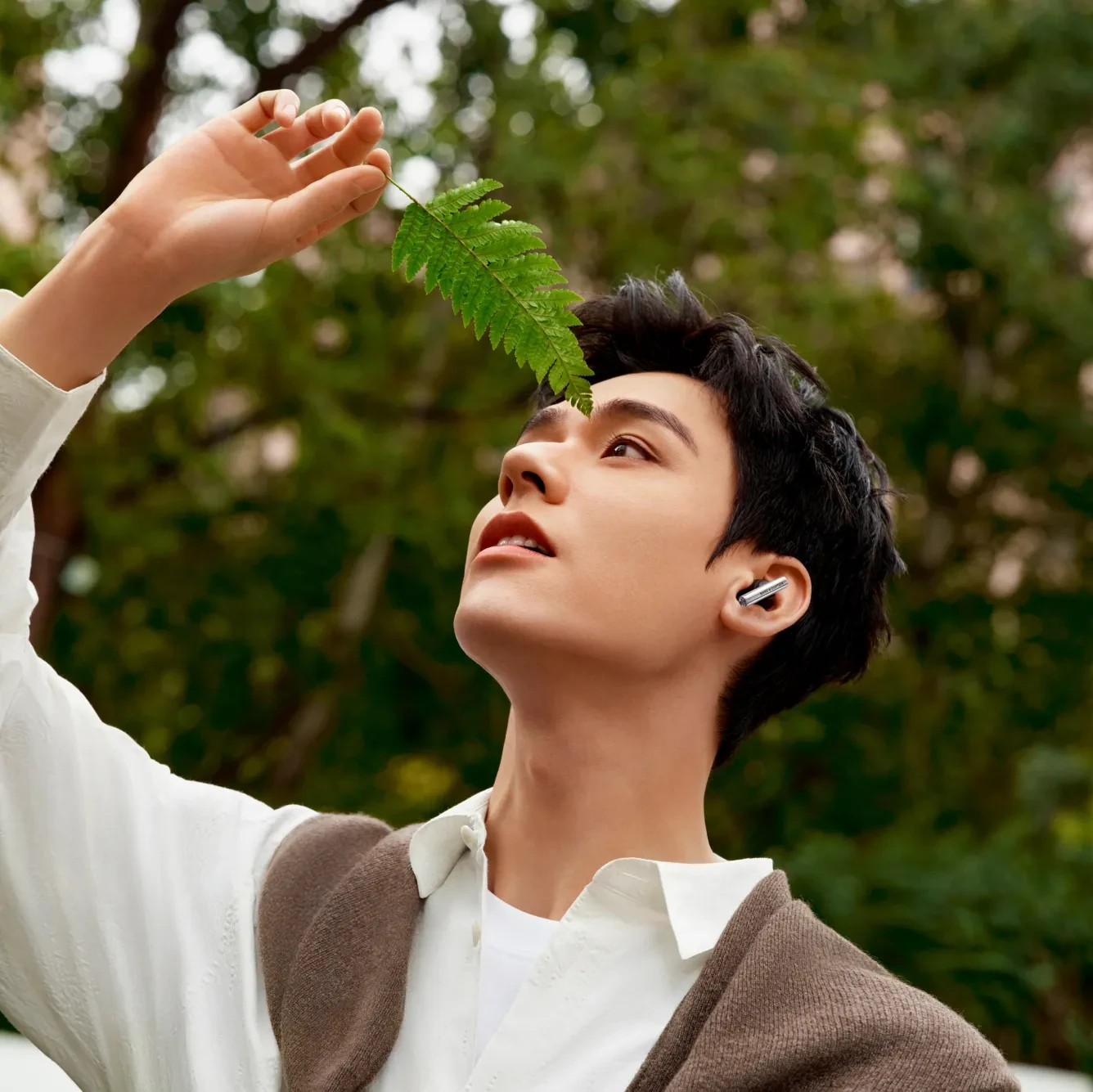 Person in white collar and brown sweater holding a green fern leaf, looking upward with wireless Beo Grace earbuds in forest setting.