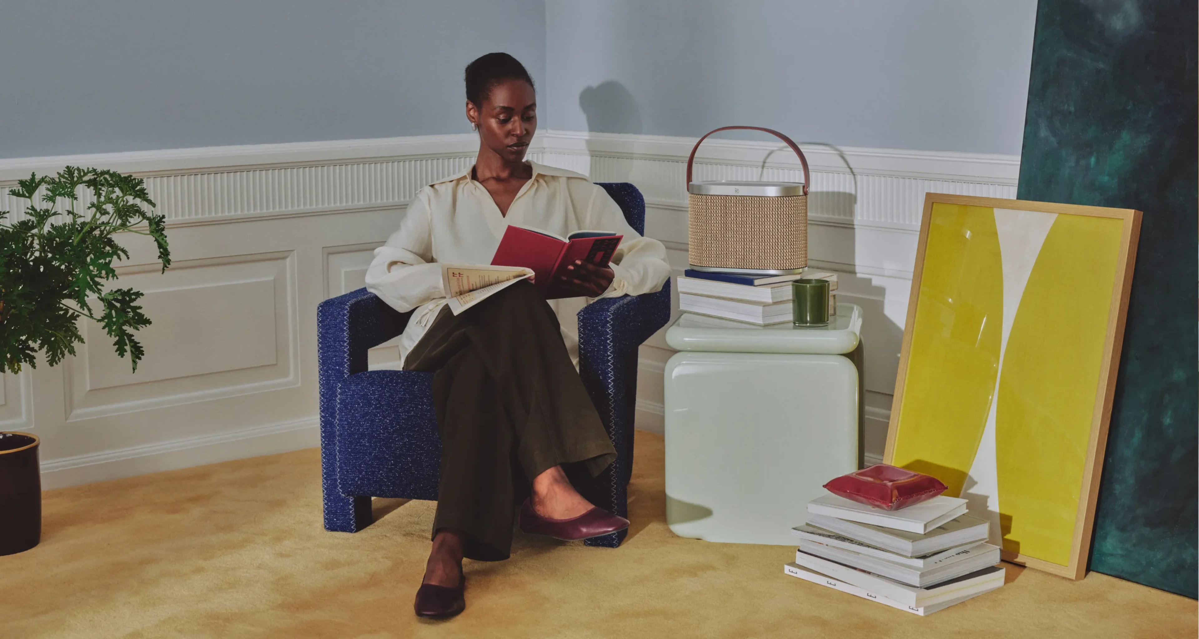 Person relaxing on an arm chair, reading a book with Beosound A5 next to them standing on a side table. Room is decorated wtith paintings and books.