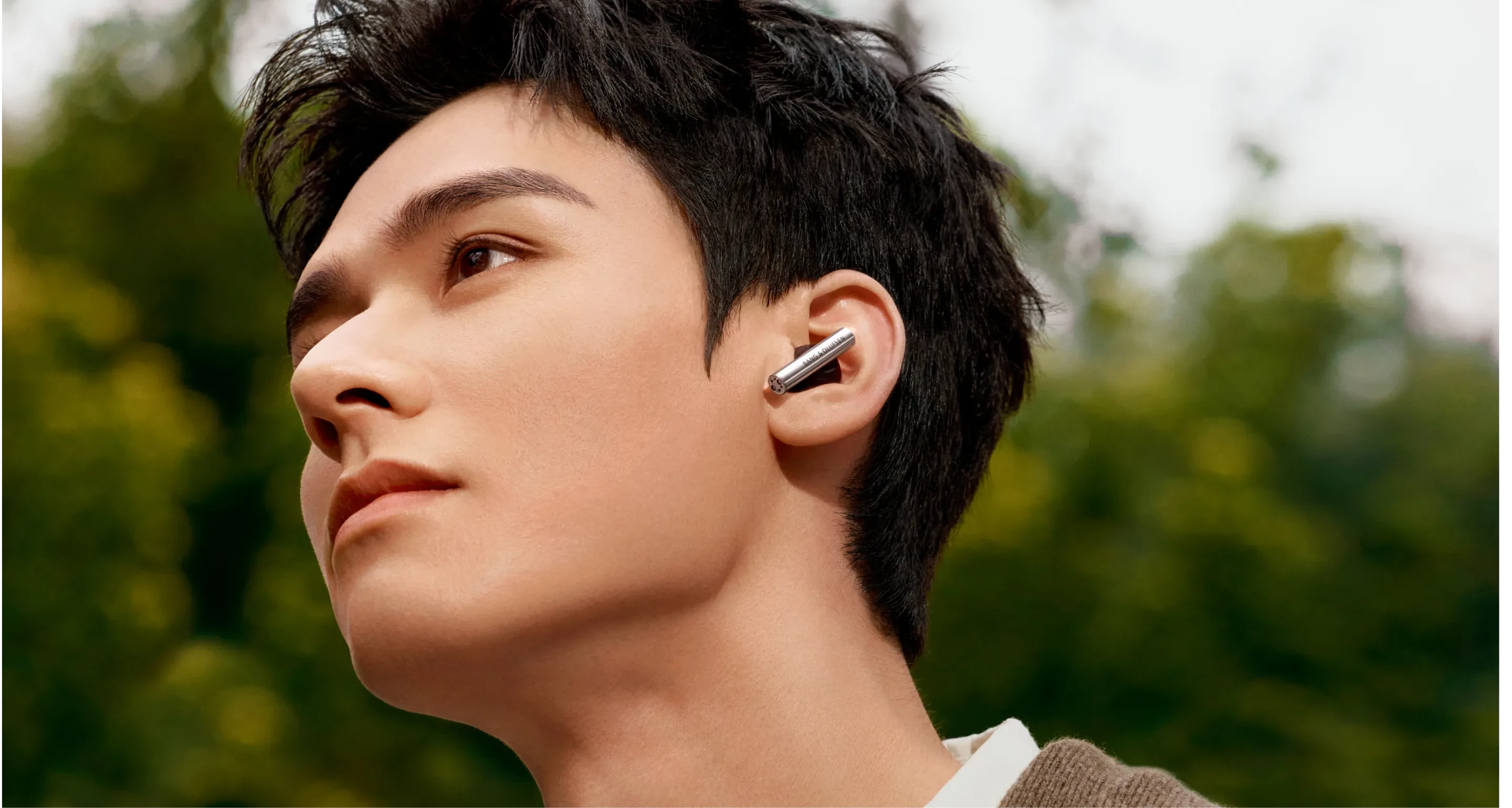 Person with short black hair wearing Beo Grace wireless earbud, photographed in profile against a blurred green background.