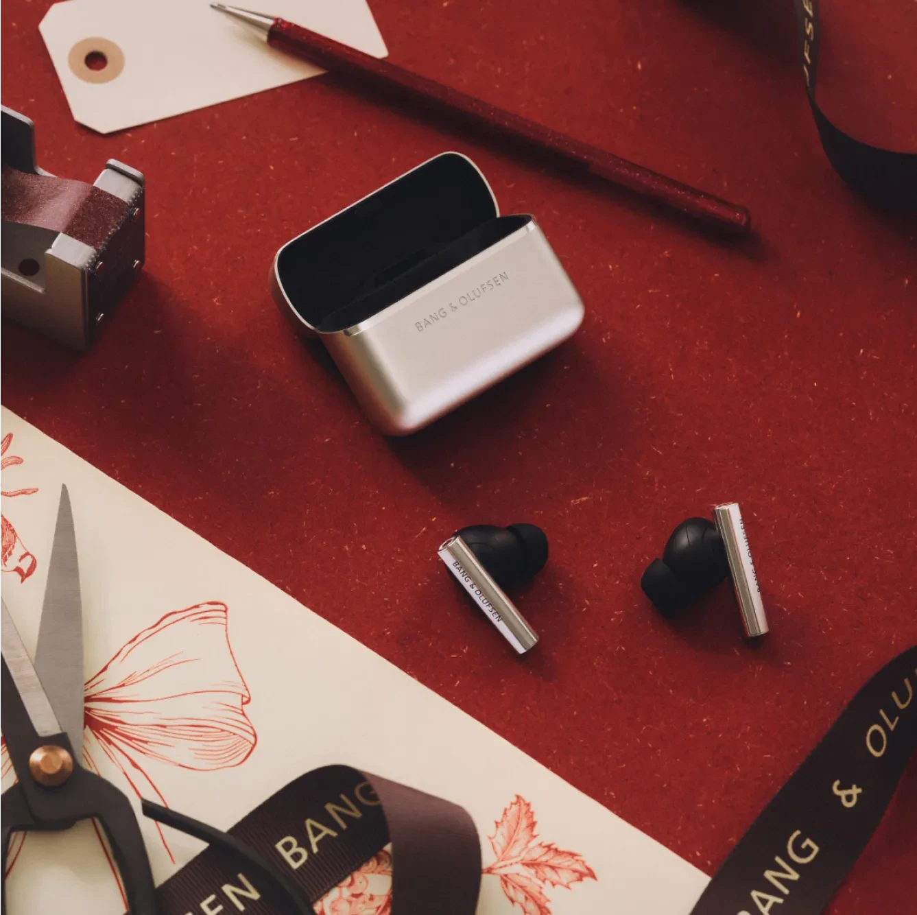 Bang & Olufsen Beo Grace wireless earbuds with charging case on red surface, surrounded by stationery and branded packaging.