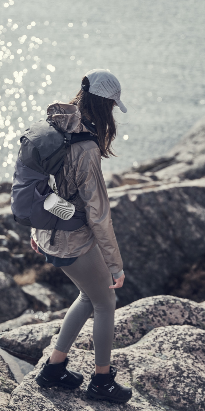 A woman hiking with Beosound Explore Grey Mist attached to her backpack with a carabiner