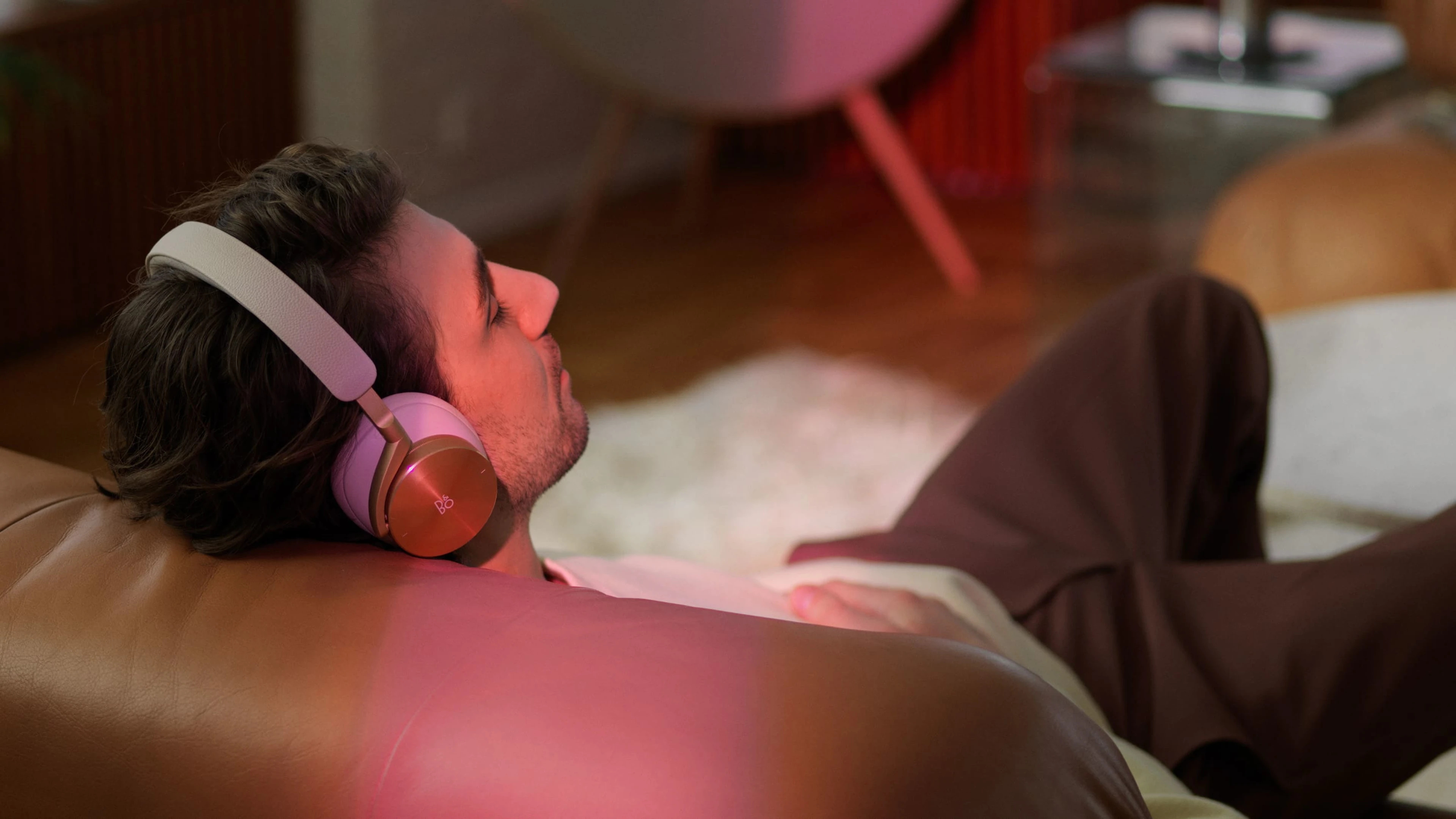 There is man seating on a sofa with closed eyes listening to music with gold tone Beoplay H95 headphones