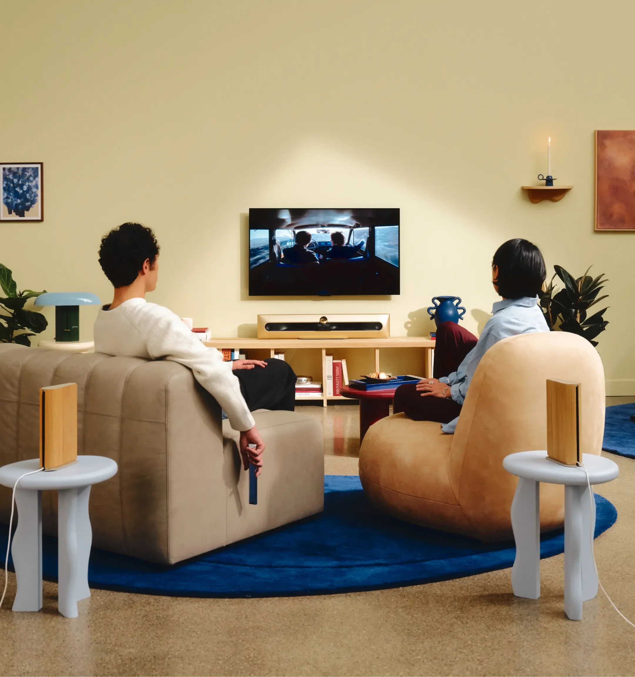 Two people watching TV with Beosound Premiere soundbar, in a stylish living room with modern furniture, blue rug, and abstract wall art.