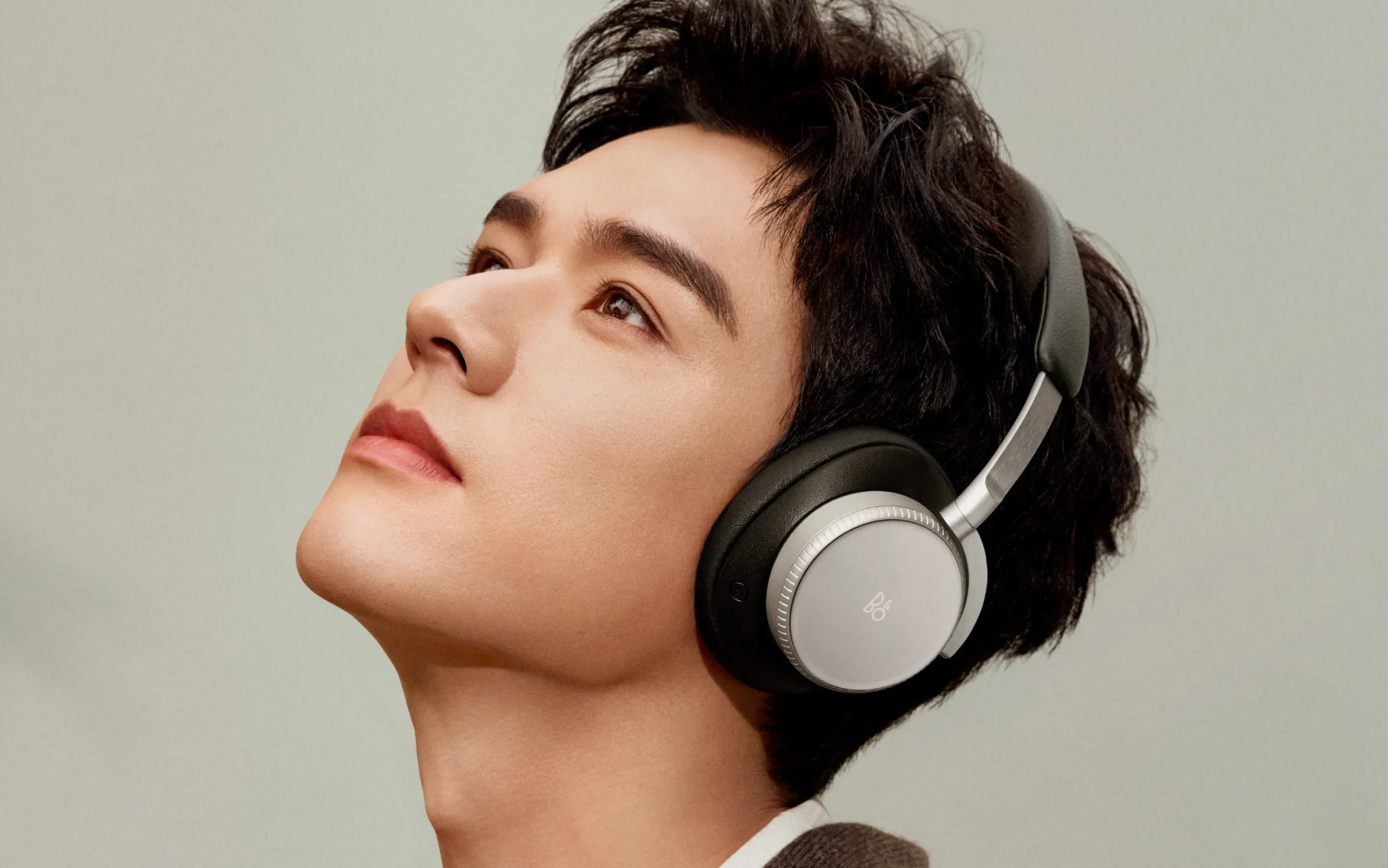 Person with dark hair wearing sleek silver and black over-ear Beoplay H100 headphones, looking upward against neutral background.