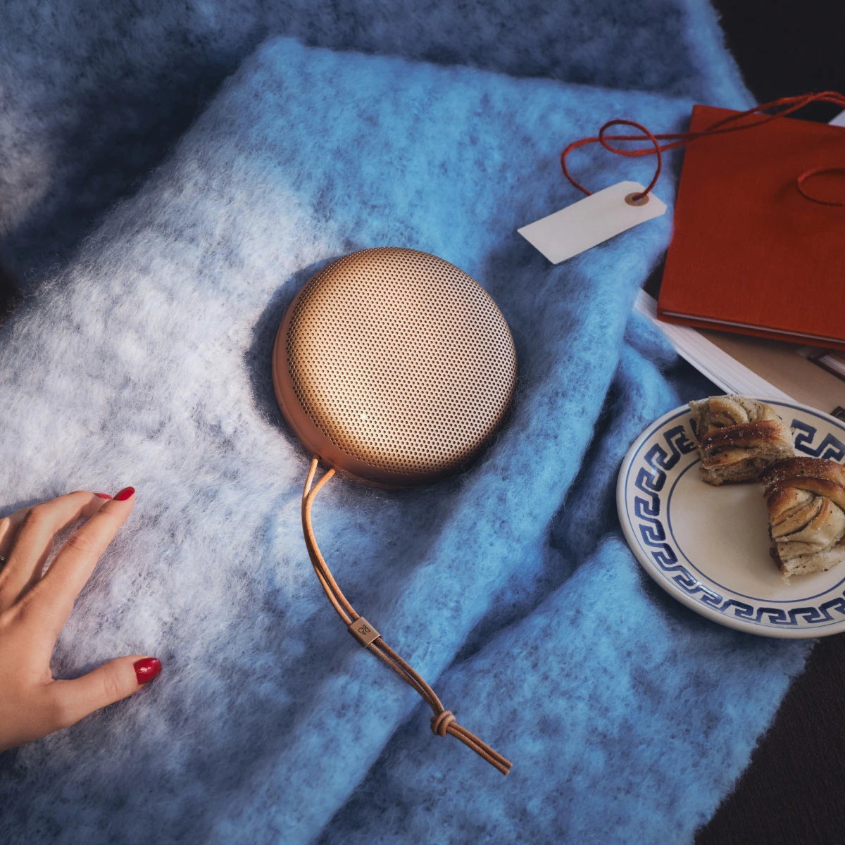 Gold portable speaker Beosound A1 on blue blanket with notebook, plate of pastry, and hand with red nails visible.