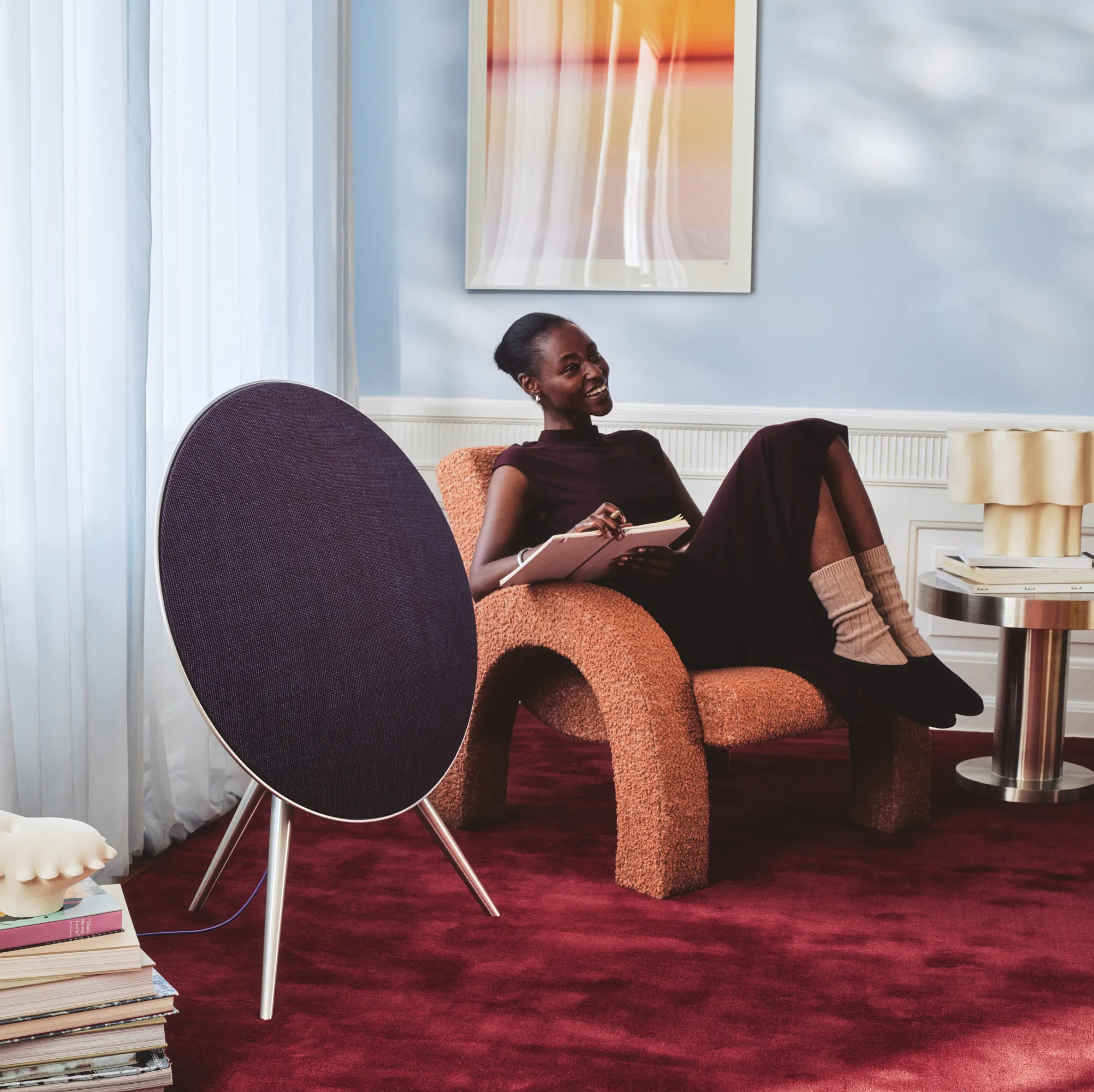 Person relaxing in orange textured chair reading a book, with circular Beoplay A9 Centennial collection speaker nearby on burgundy carpet in stylish room.