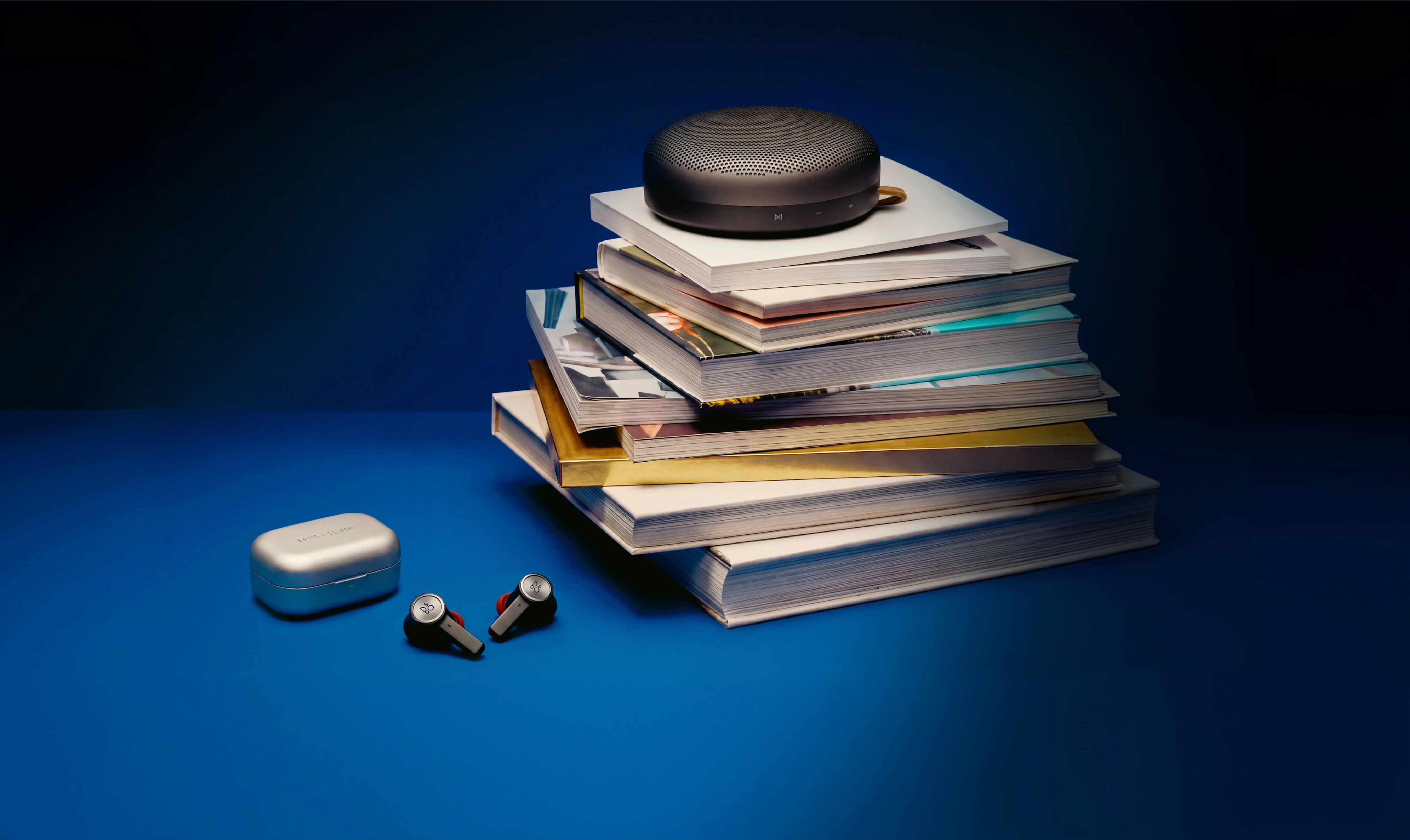 Warm Granite 색상의 Beosound A1이 책 더미 위에 놓여 있고, 그 옆에는 Beoplay Eleven 이어버드가 꺼내진 상태로 함께 놓여 있습니다.