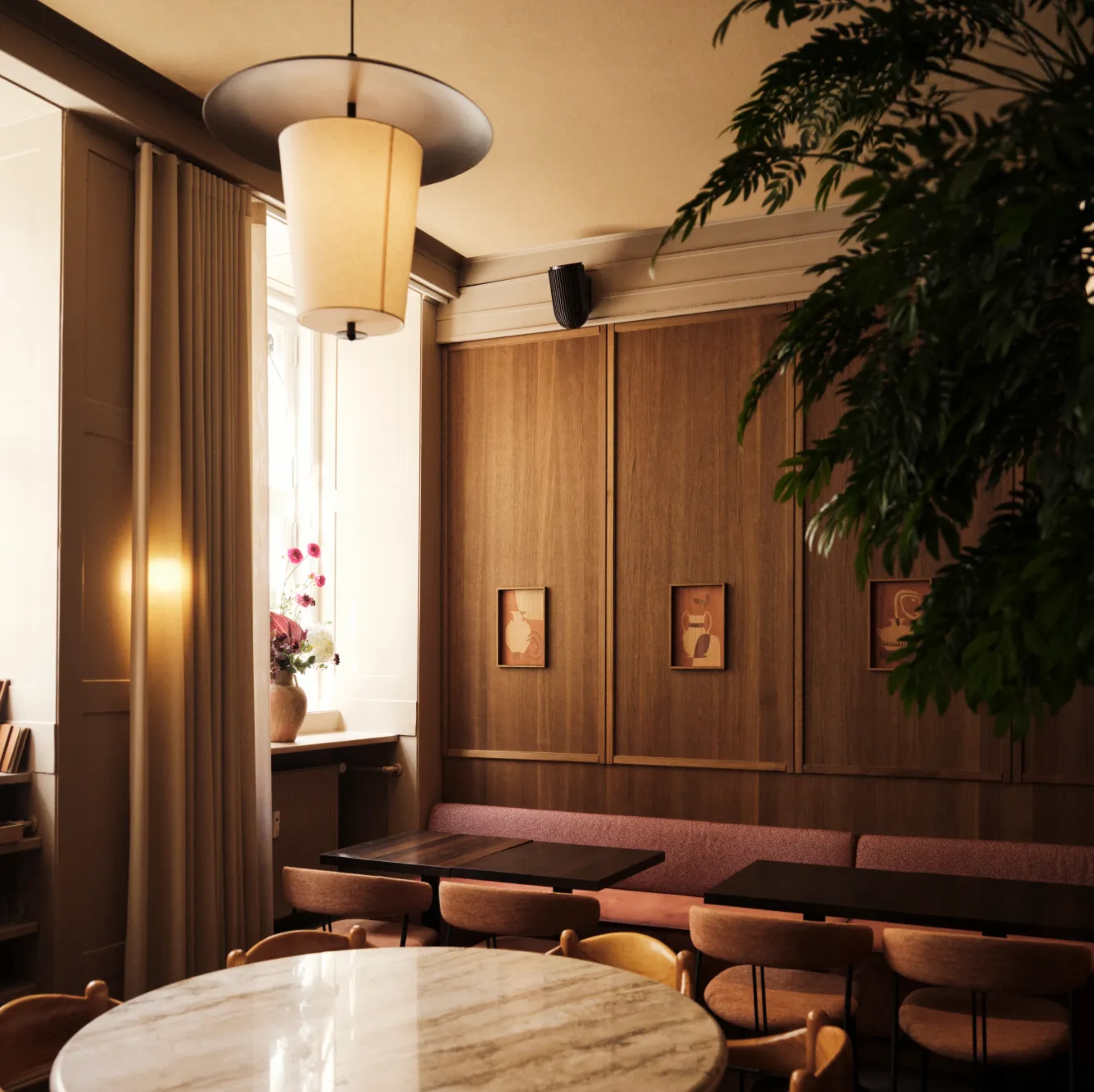 Warm, wood-paneled dining interior with upholstered banquette seating, a large round marble table, and a sculptural pendant light suspended above.