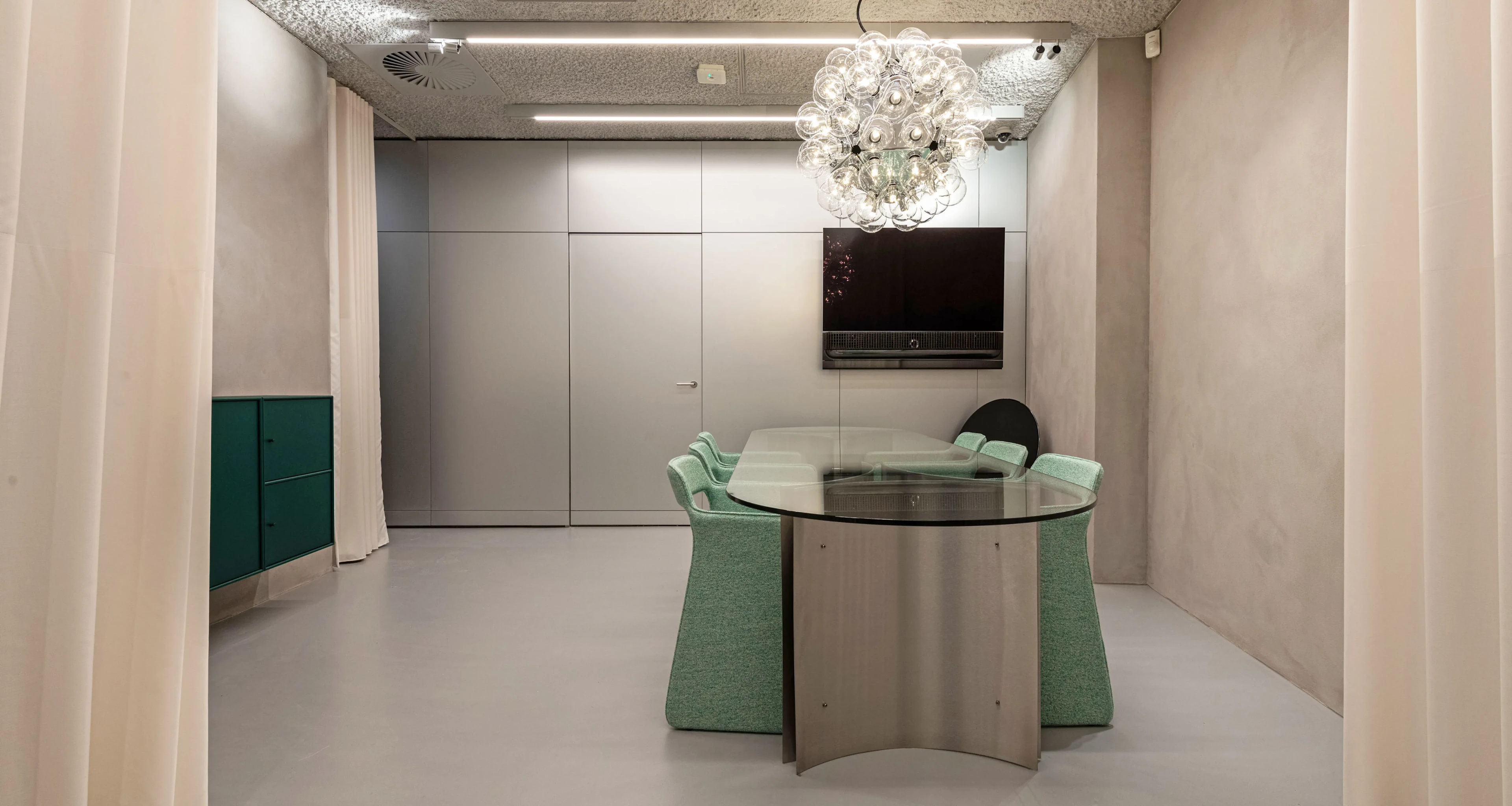 Meeting room in our stores with round glass table, mint green chairs, bubble chandelier, and wall-mounted screen.