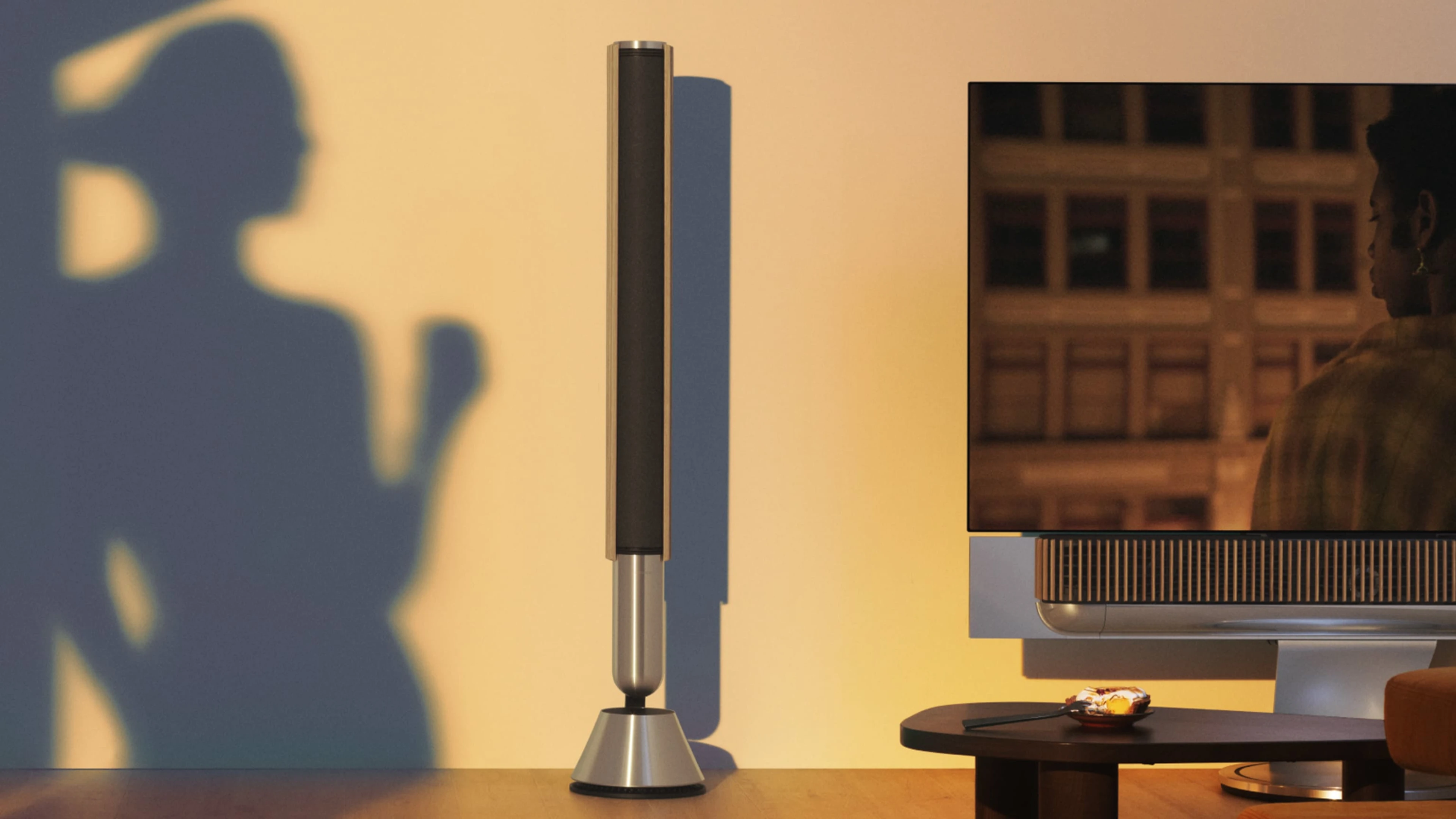 Beolab 28 in natural aluminum and light oak next to a tv in a living room with a woman's shadow