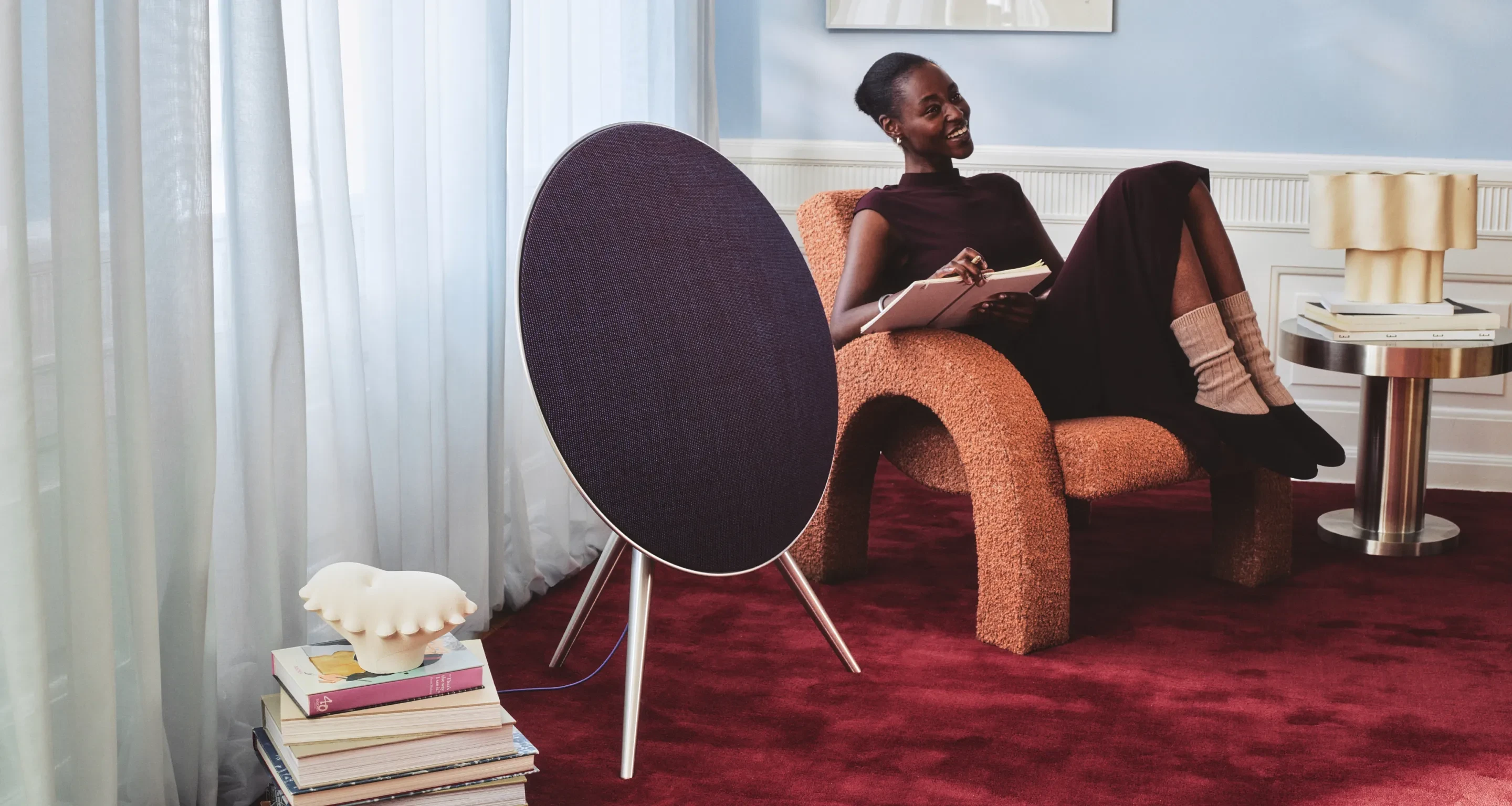 Beosound A9 in the variant Century Blue in a cozy room with a woman sitting next to the speaker, writing in her journal.