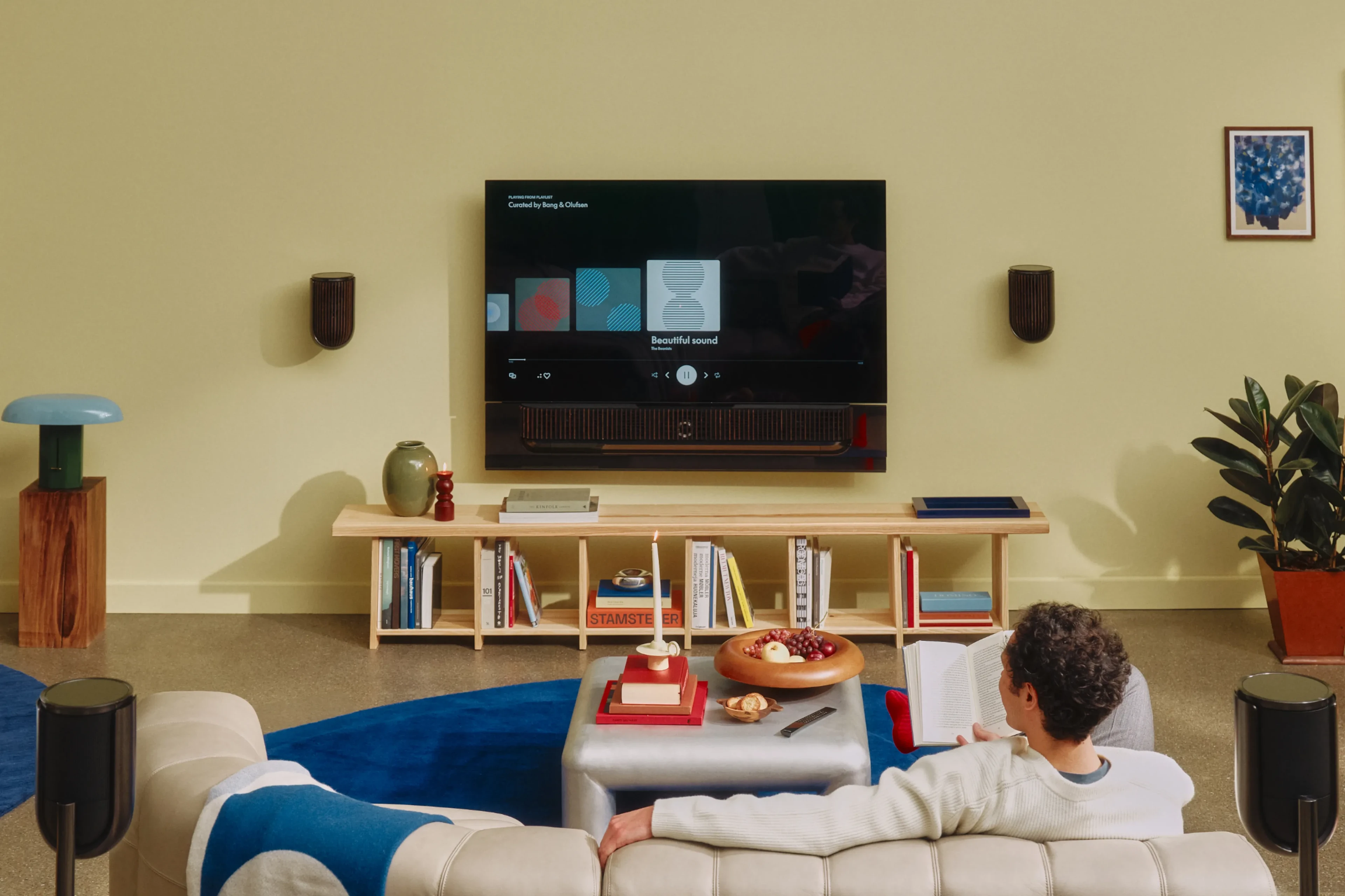 A guy sitting on a sofa in a modern interior living room watching TV, and there is Beosound Theatre soundbar below the TV, and a set of Beolab 8 next to the TV attached to the wall
