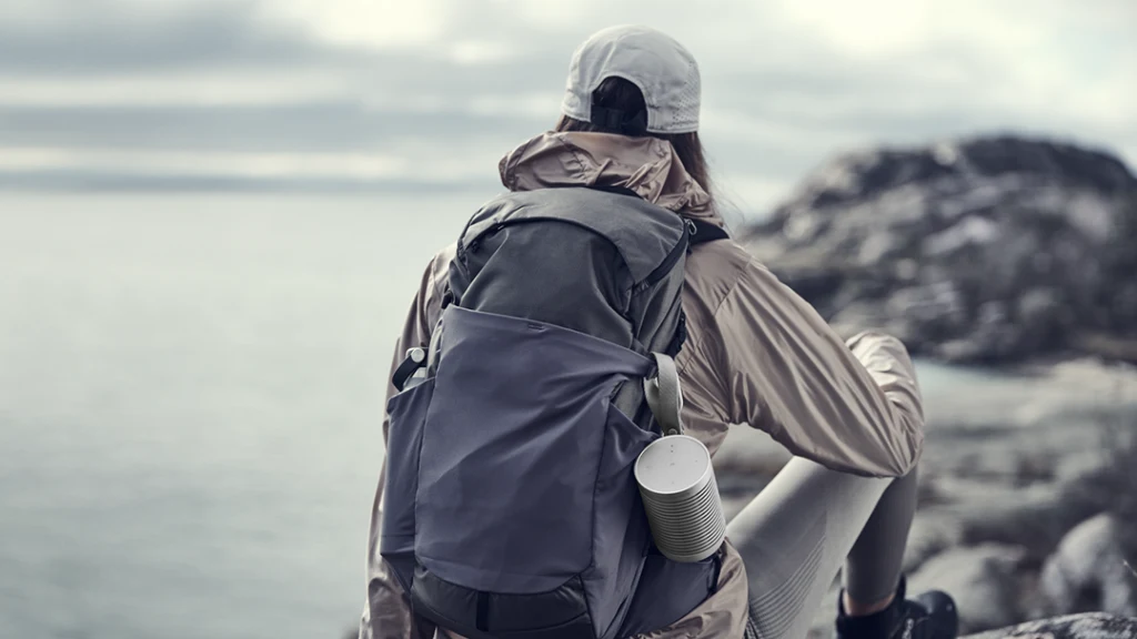 Beosound Explore hangs from a woman's backpack while she is resting sitting on a rock