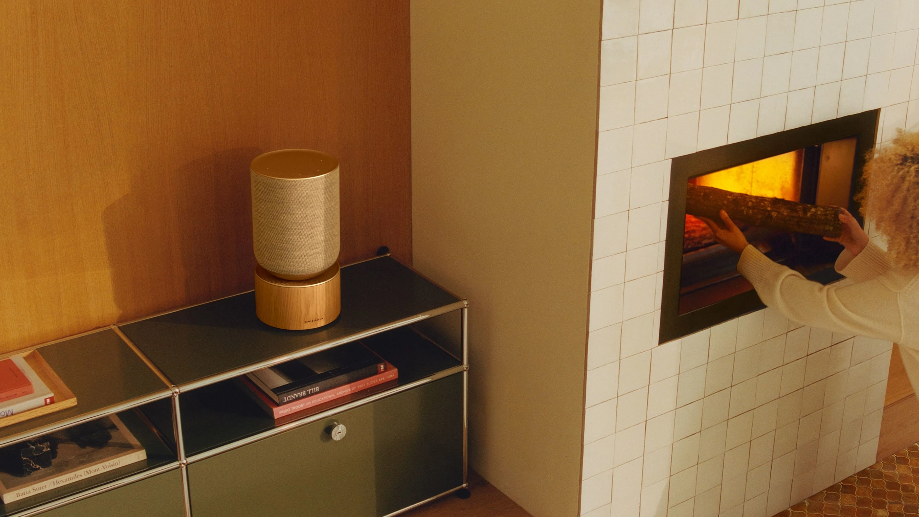 Beosound Balance in natural oak placed on a green modular furniture next to a fireplace where the lady is putting the wood in
