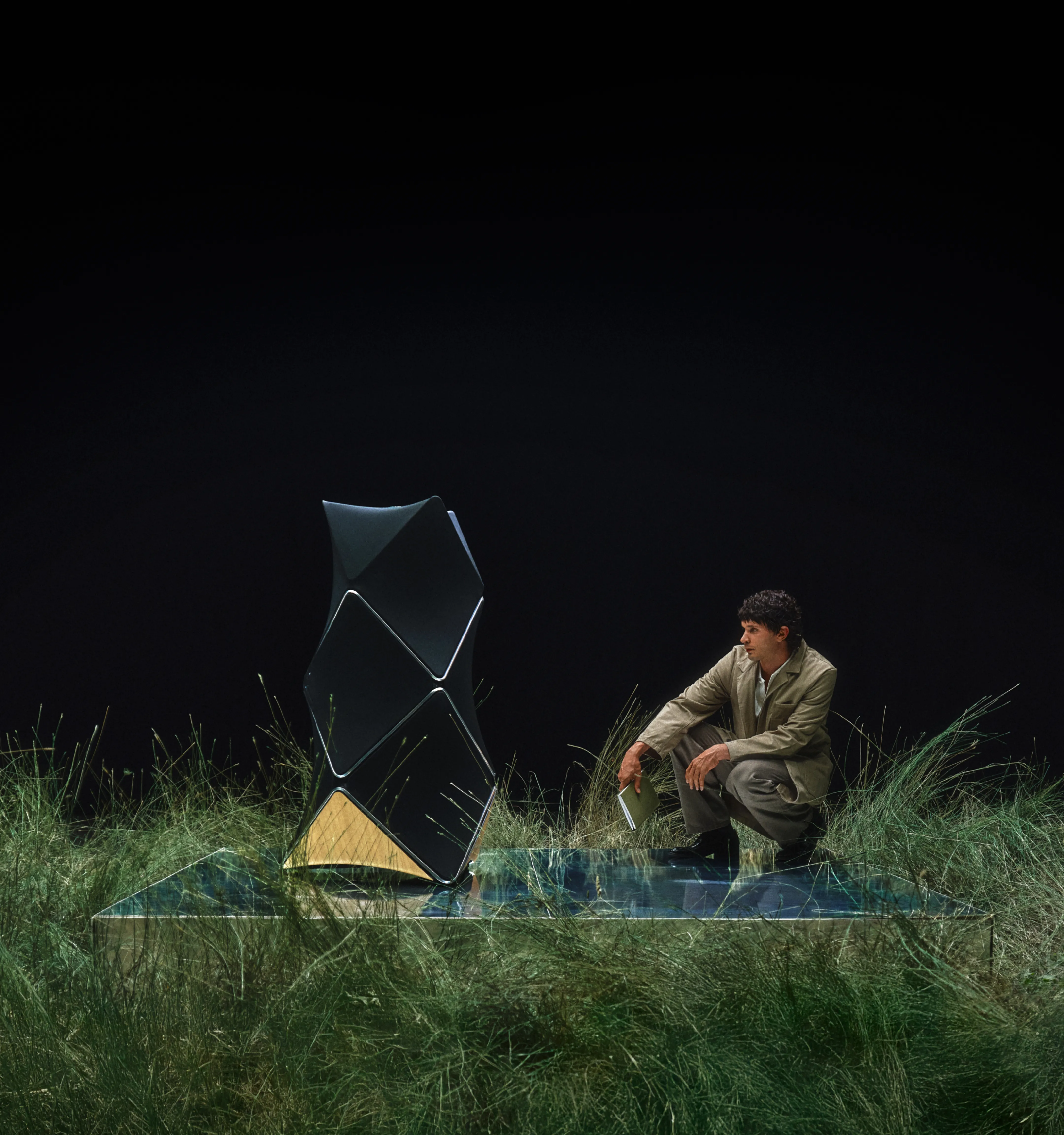 Person in beige outfit sitting on glass platform with Beolab 90 and sheep amid tall grass against black background.
