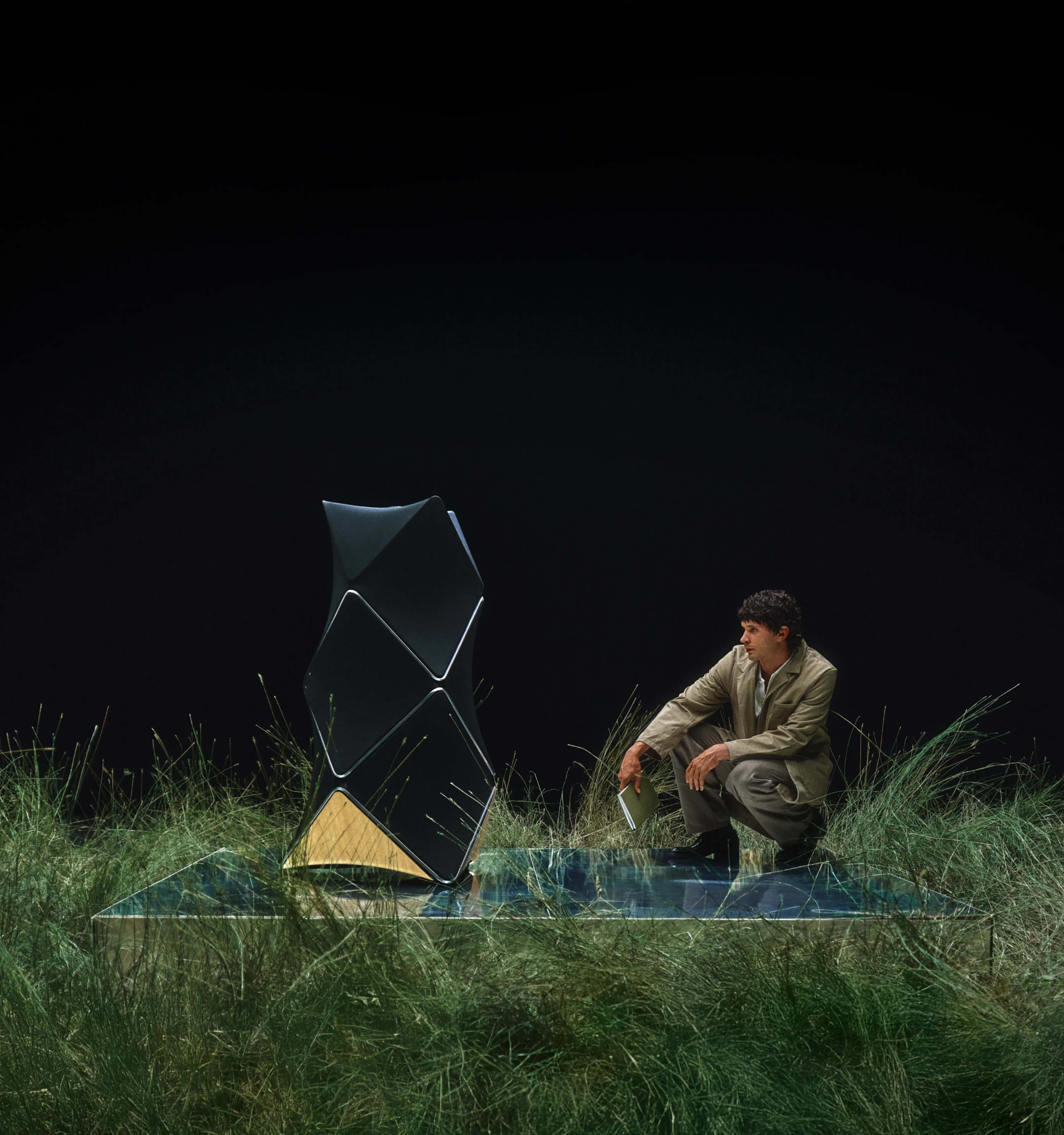 Person in beige outfit sitting on glass platform with Beolab 90 and sheep amid tall grass against black background.