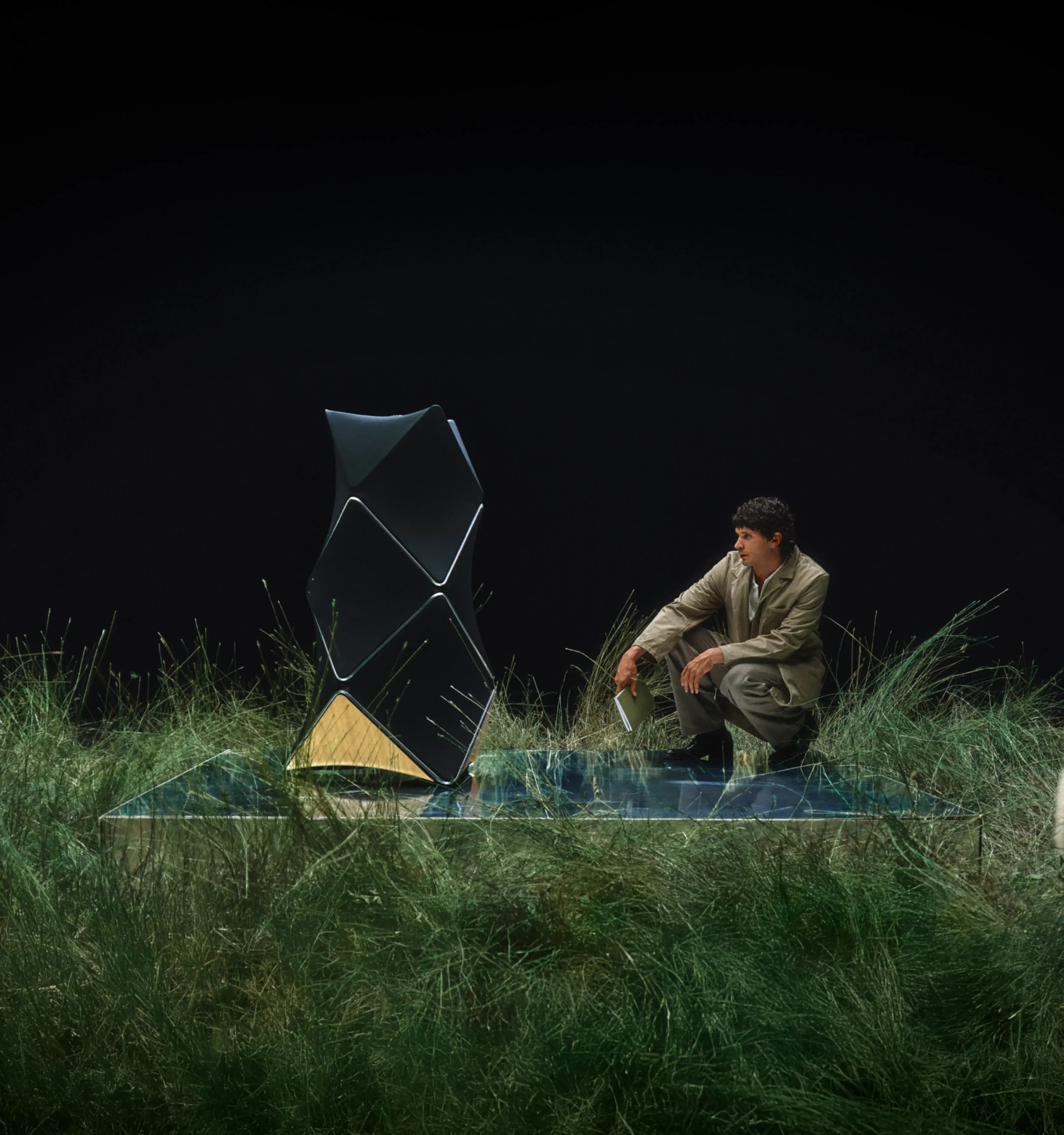 Person in beige outfit sitting on glass platform with Beolab 90 and sheep amid tall grass against black background.