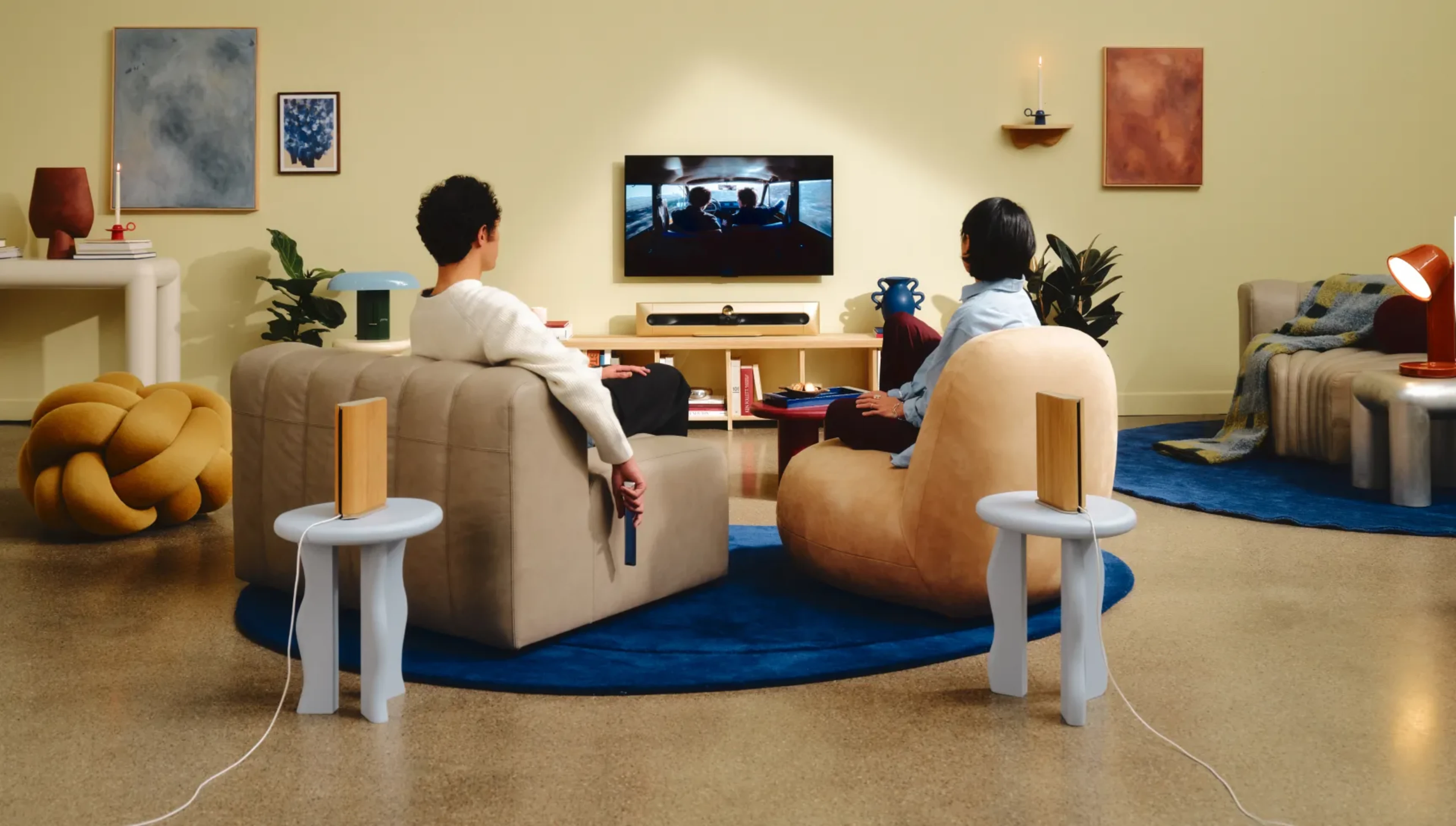 Two people watching TV with Beosound Premiere soundbar, in a stylish living room with modern furniture, blue rug, and abstract wall art.