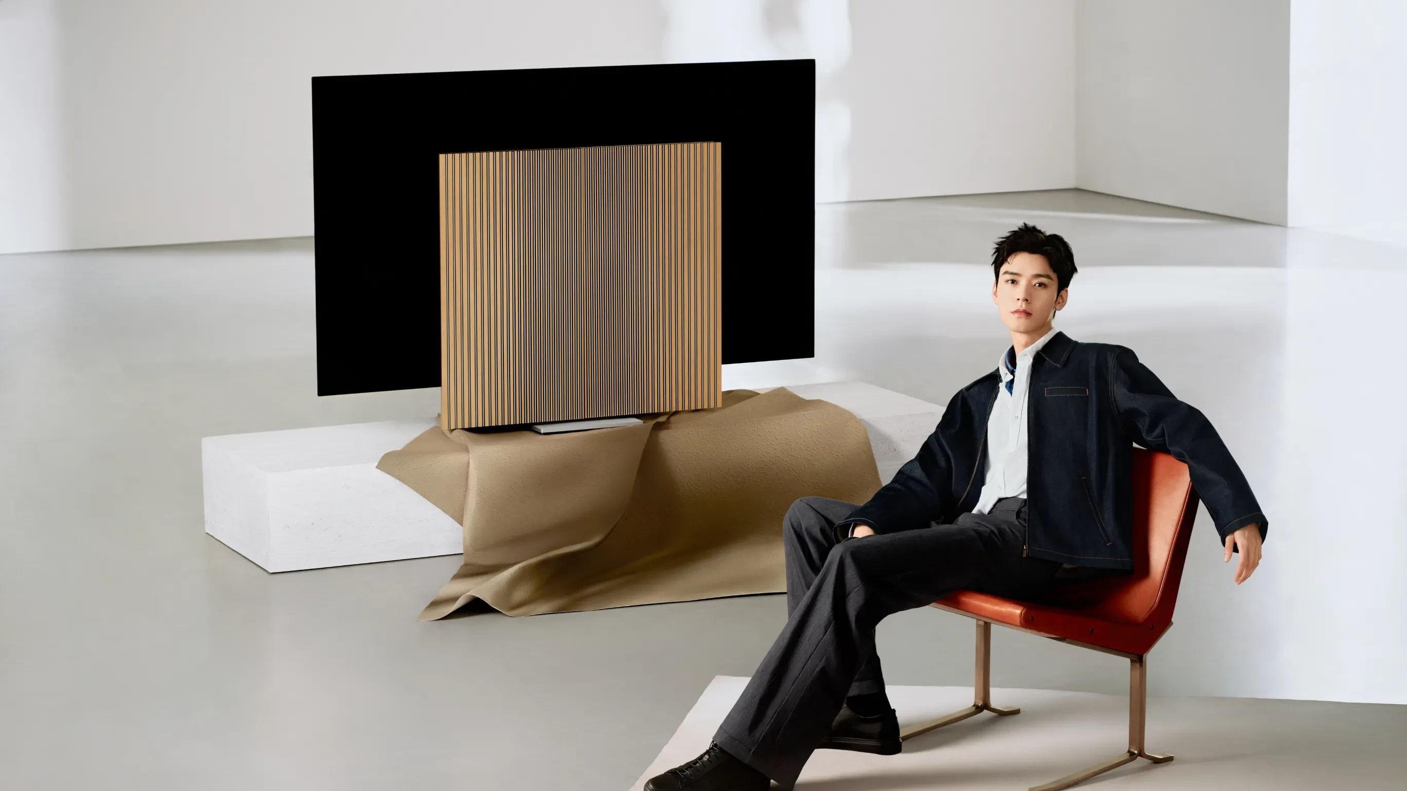Person in dark outfit seated on red chair beside Beovision Harmony television with wooden ribbed front panel in minimalist white room.