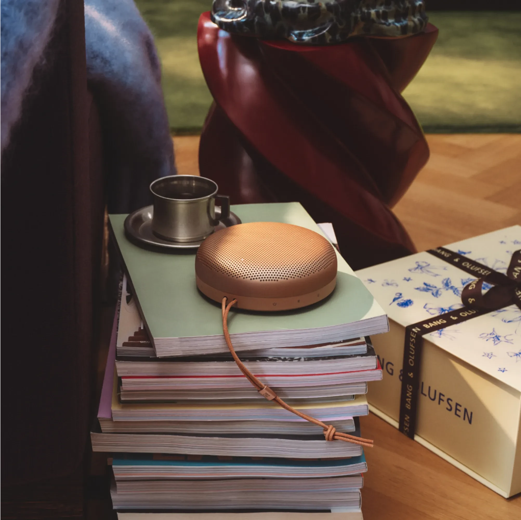 Copper Beosound A1 portable speaker on stack of magazines with metal coffee cup and red chair in cozy reading corner.
