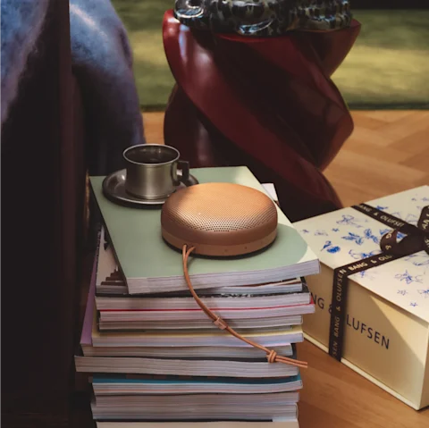 Copper Beosound A1 portable speaker on stack of magazines with metal coffee cup and red chair in cozy reading corner.