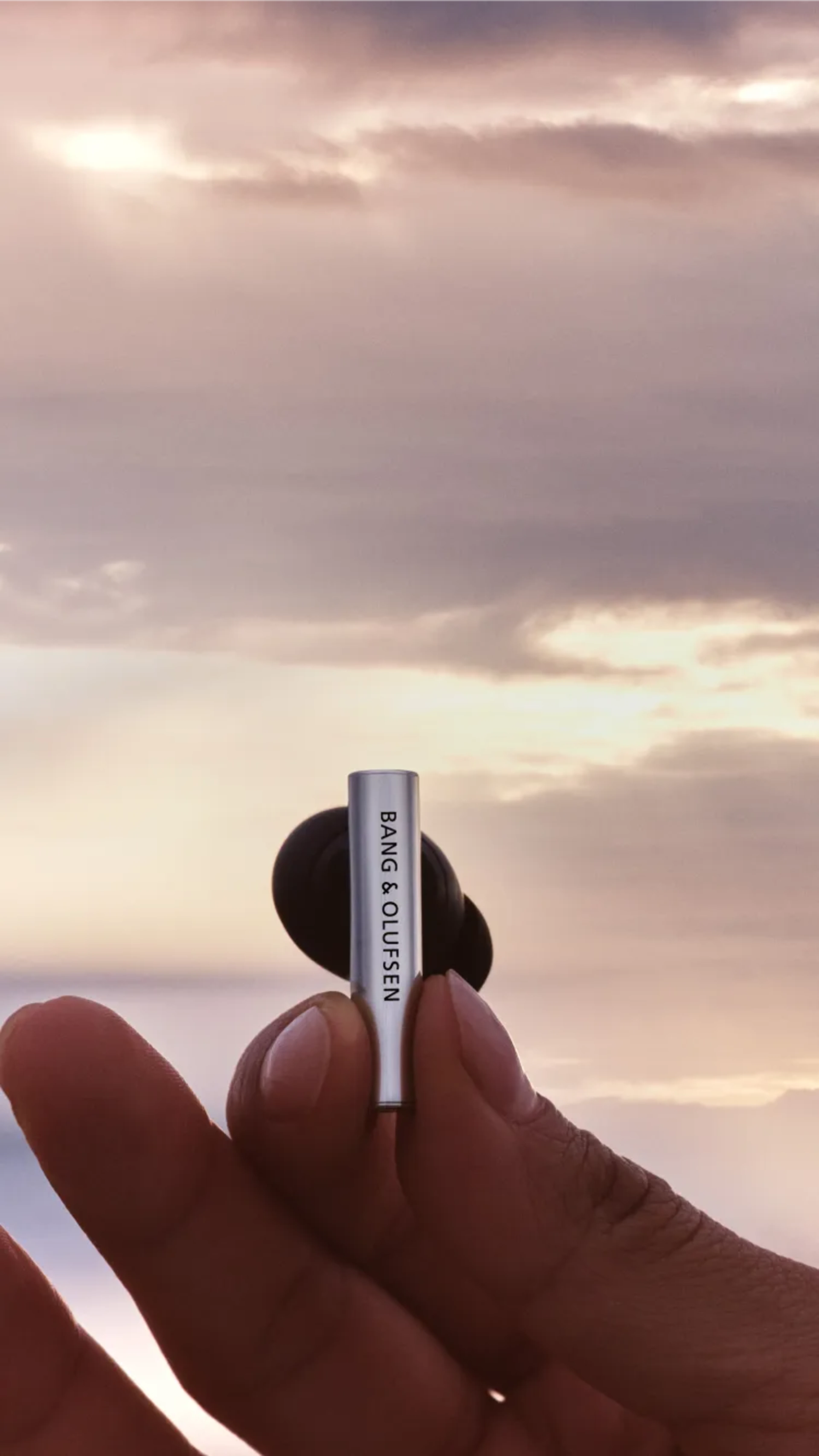 一隻手握著 Beo Grace 入耳式耳機,背後則是一幅美麗的日落背景