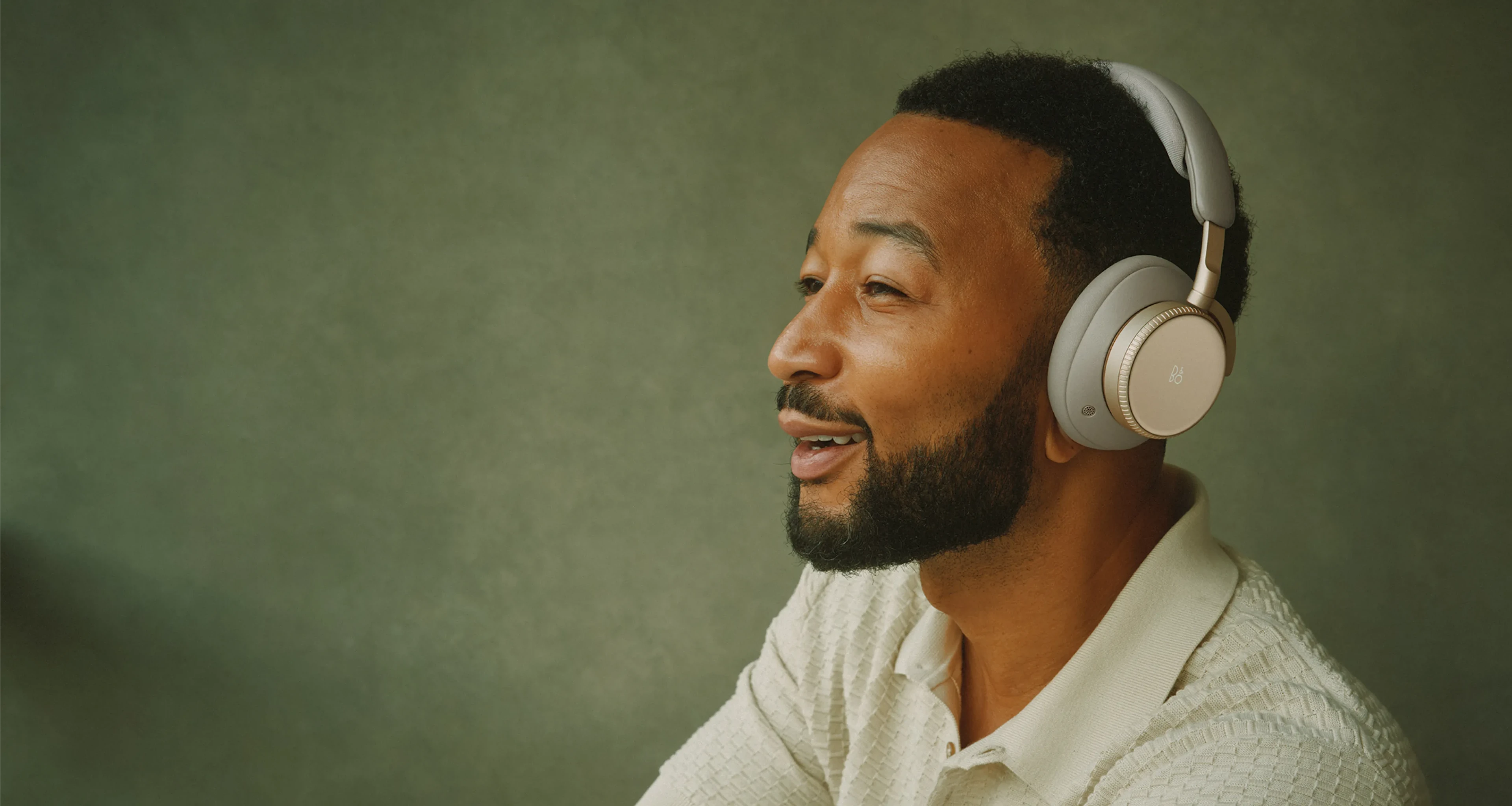 John Legend depicted wearing Bang & Olufsen Beoplay H100 over-ear headphones in the Hourglass Sand colorway, photographed against a green background in a relaxed, music-listening pose.
