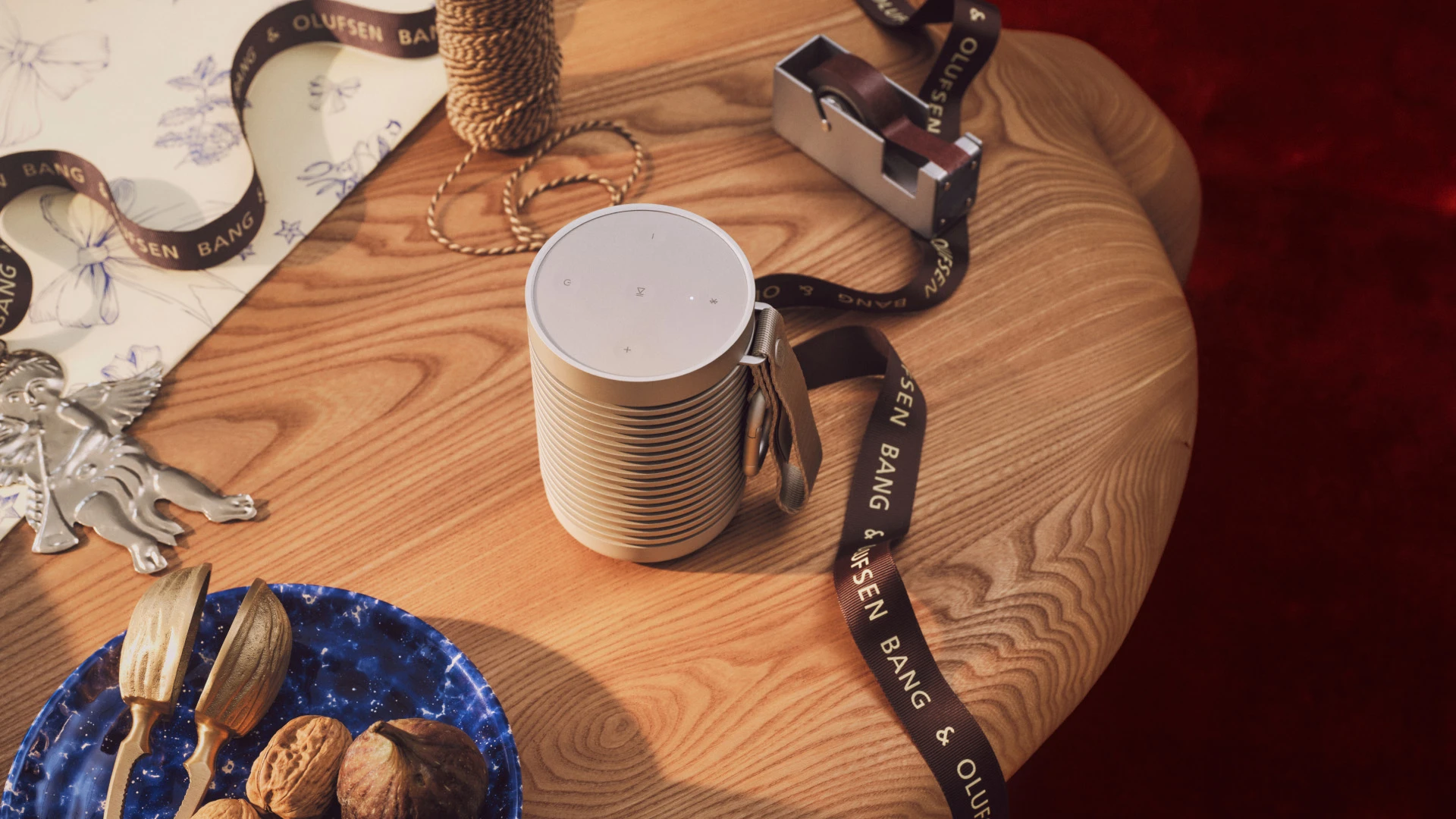 Beosound Explore Bang & Olufsen speaker on wooden table with branded ribbon, twine, and blue plate with walnuts.