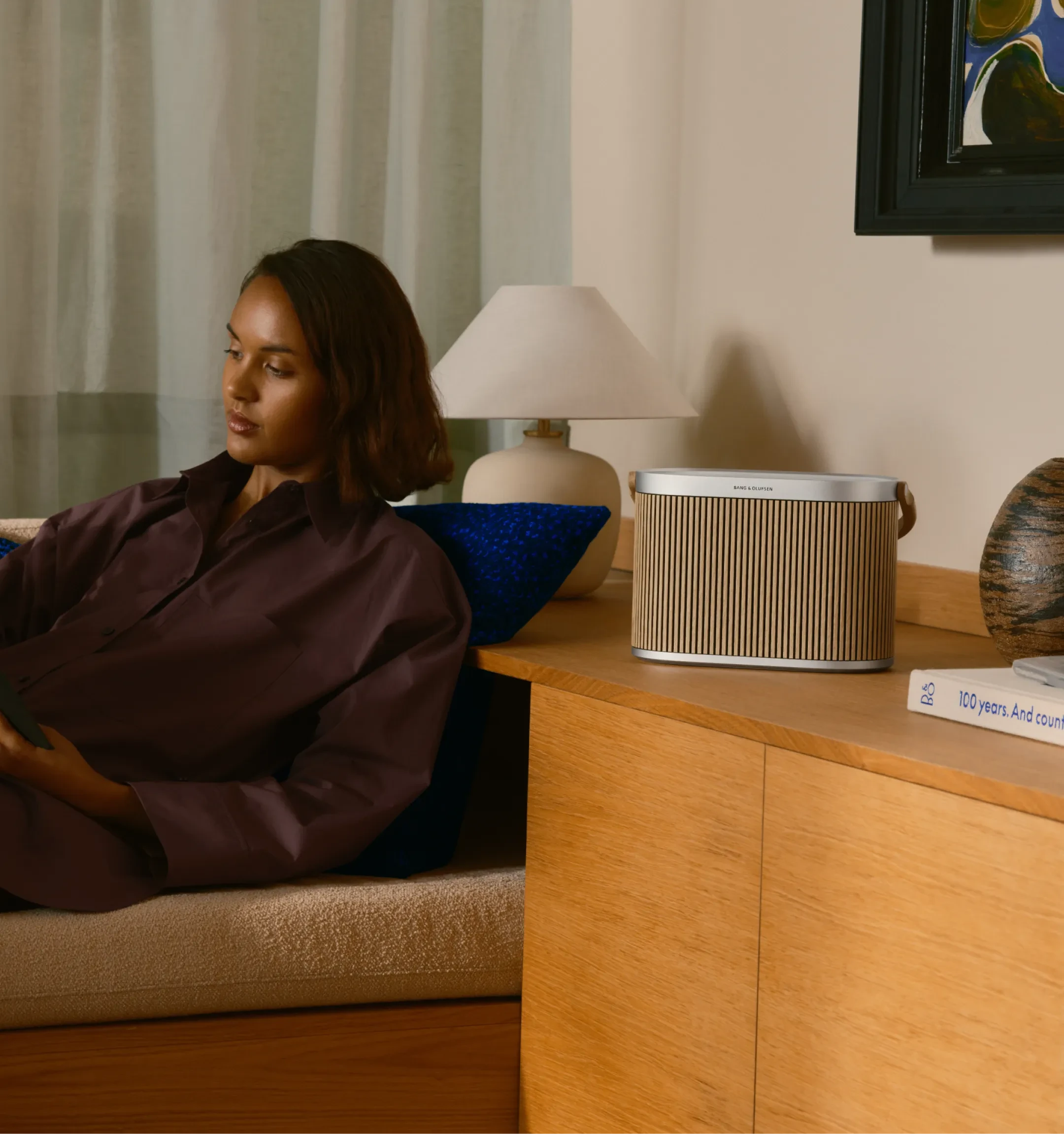 A woman in a brown shirt, lounging on a sofa with Beosound A5 on a side table.