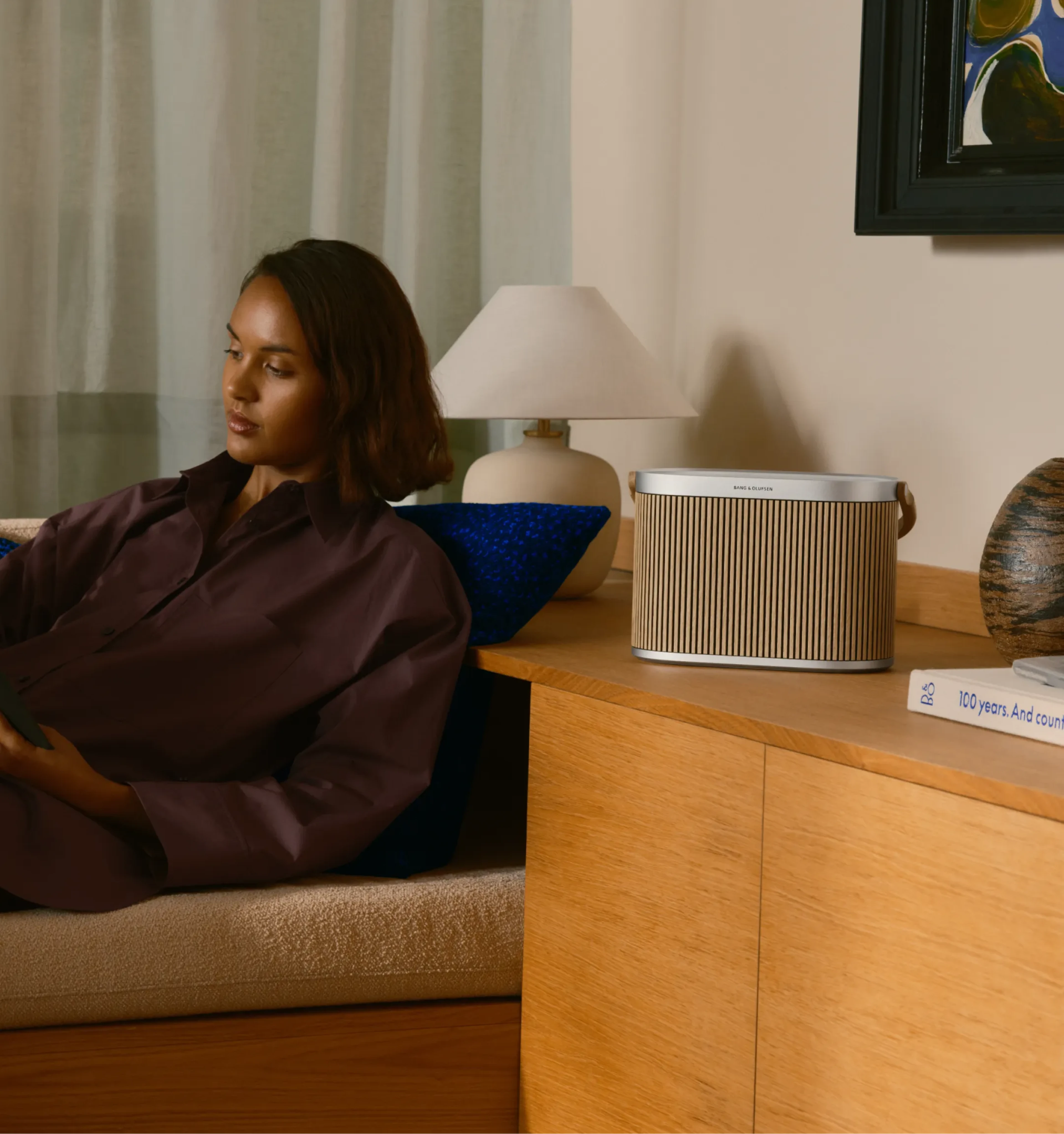A woman in a brown shirt, lounging on a sofa with Beosound A5 on a side table.