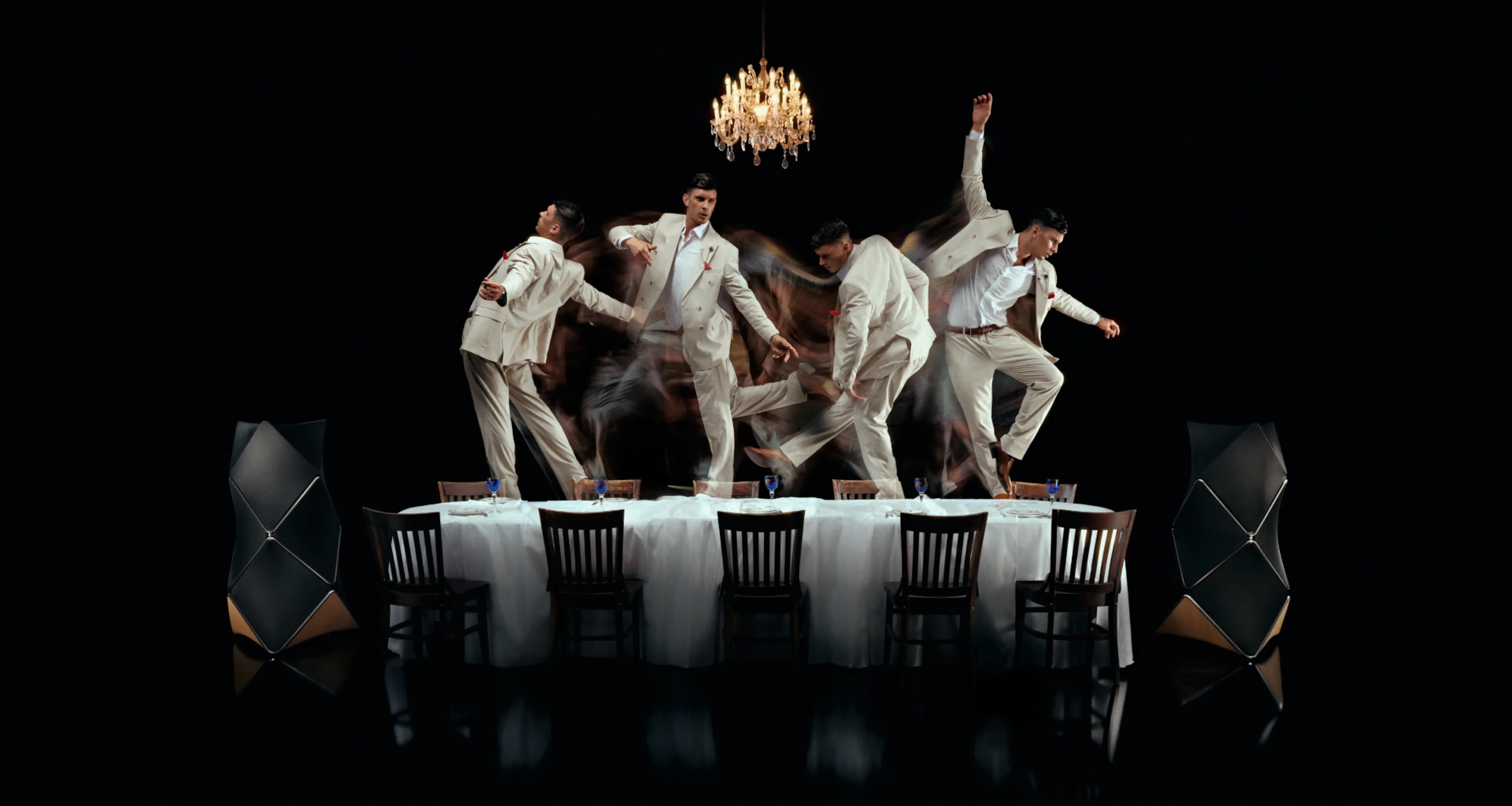 Dancers in white suits performing on a dining table under a crystal chandelier, creating motion blur against a dark background.