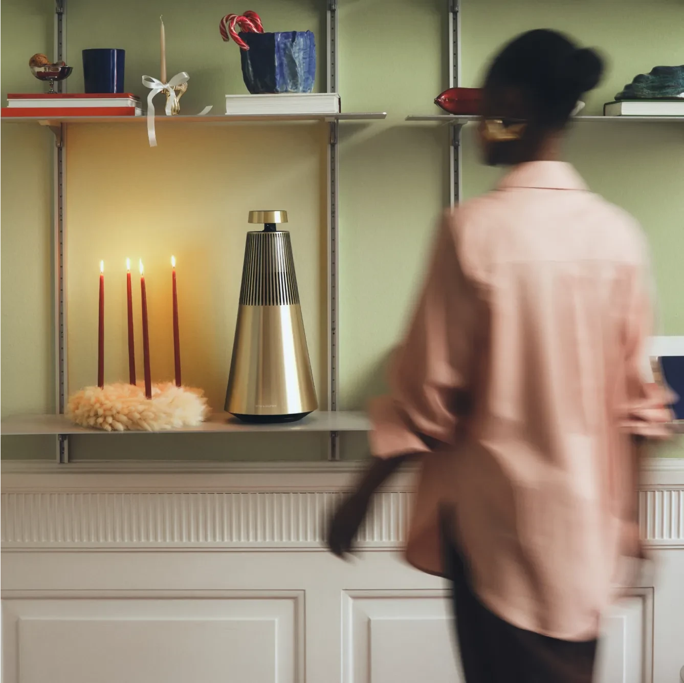 Blurred figure in pink passing shelves with lit candles, a metallic Beosound 2 speaker, books, and decorative items against green wall.