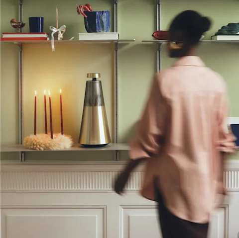 Blurred figure in pink passing shelves with lit candles, a metallic Beosound 2 speaker, books, and decorative items against green wall.