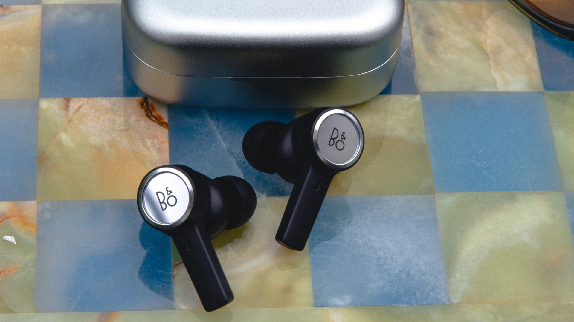 Natural aluminium Beoplay Eleven earbuds on a colorful check pattern desk