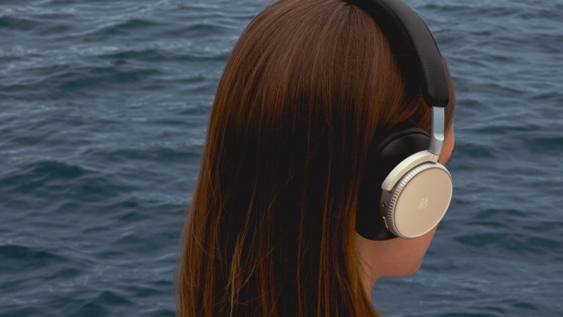 A woman wearing black Beoplay H100 by the water