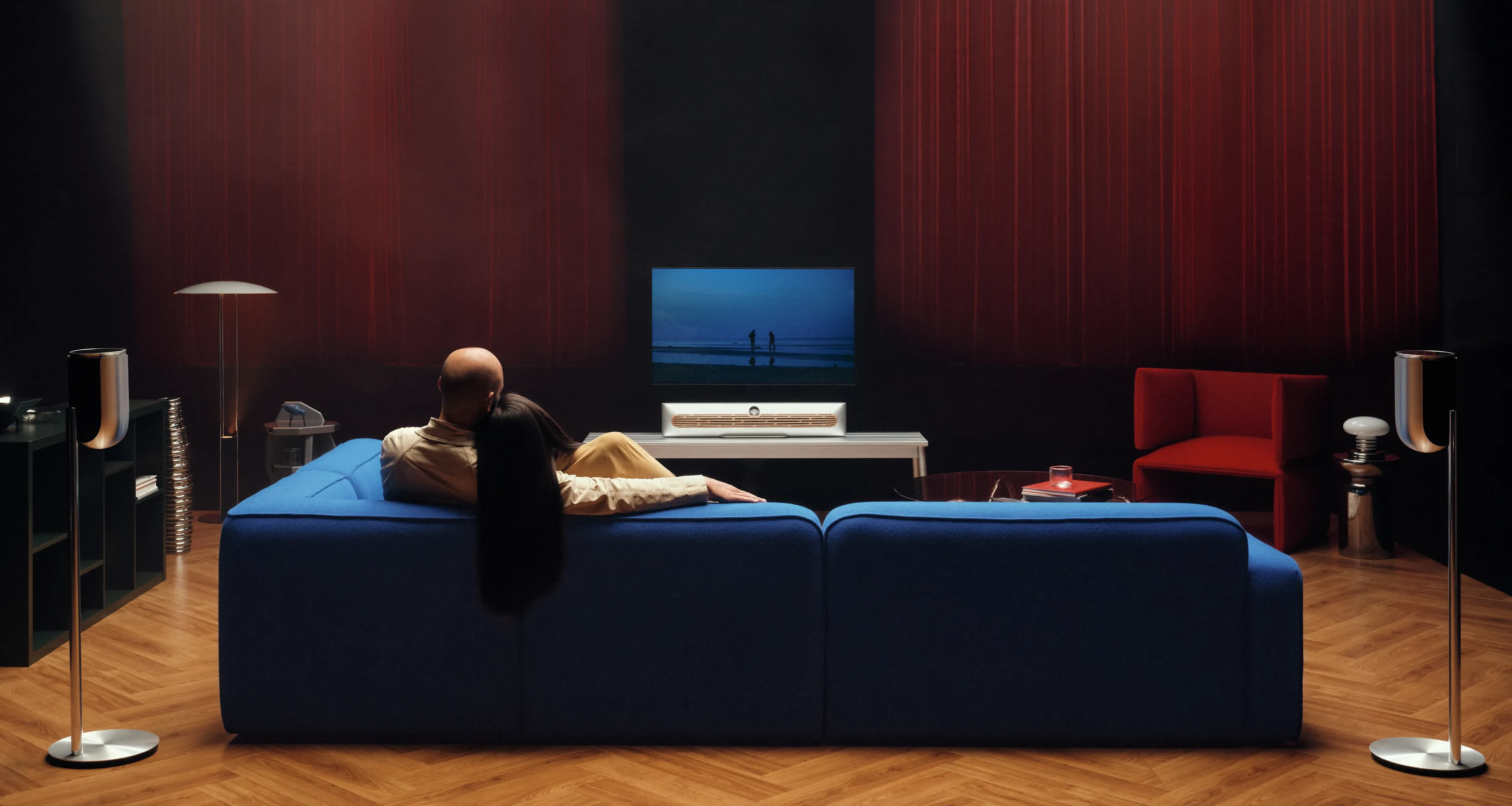 一对夫妇坐在沙发上看电视,搭配 Beosound Premiere 扬声器,沙发后面两边分别放着一对 Beolab 8