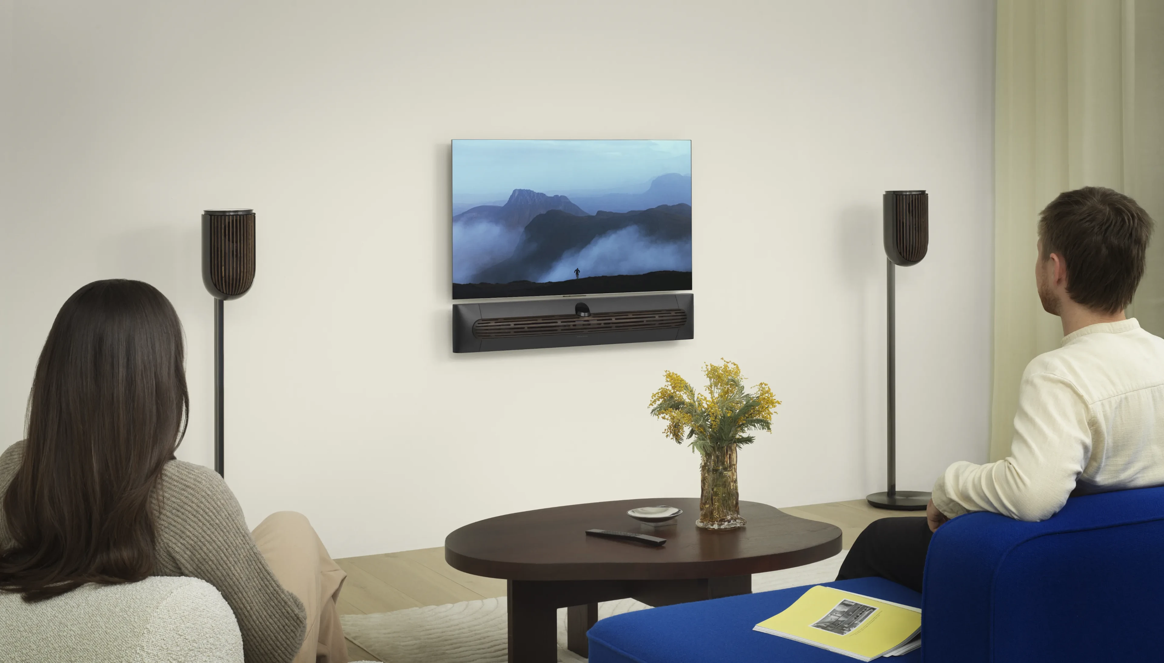 A couple sitting on separate sofas watching TV and there is Beosound Premiere under the TV and a set of Beolab 8 on each side