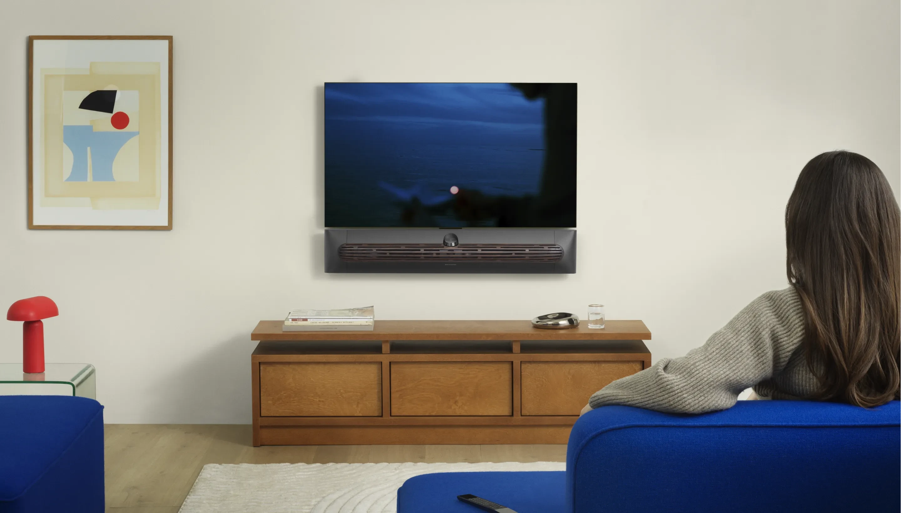 Beosound Premiere Black Anthracite in a living room, mounted on the wall with a TV, with a person sitting on a blue sofa.