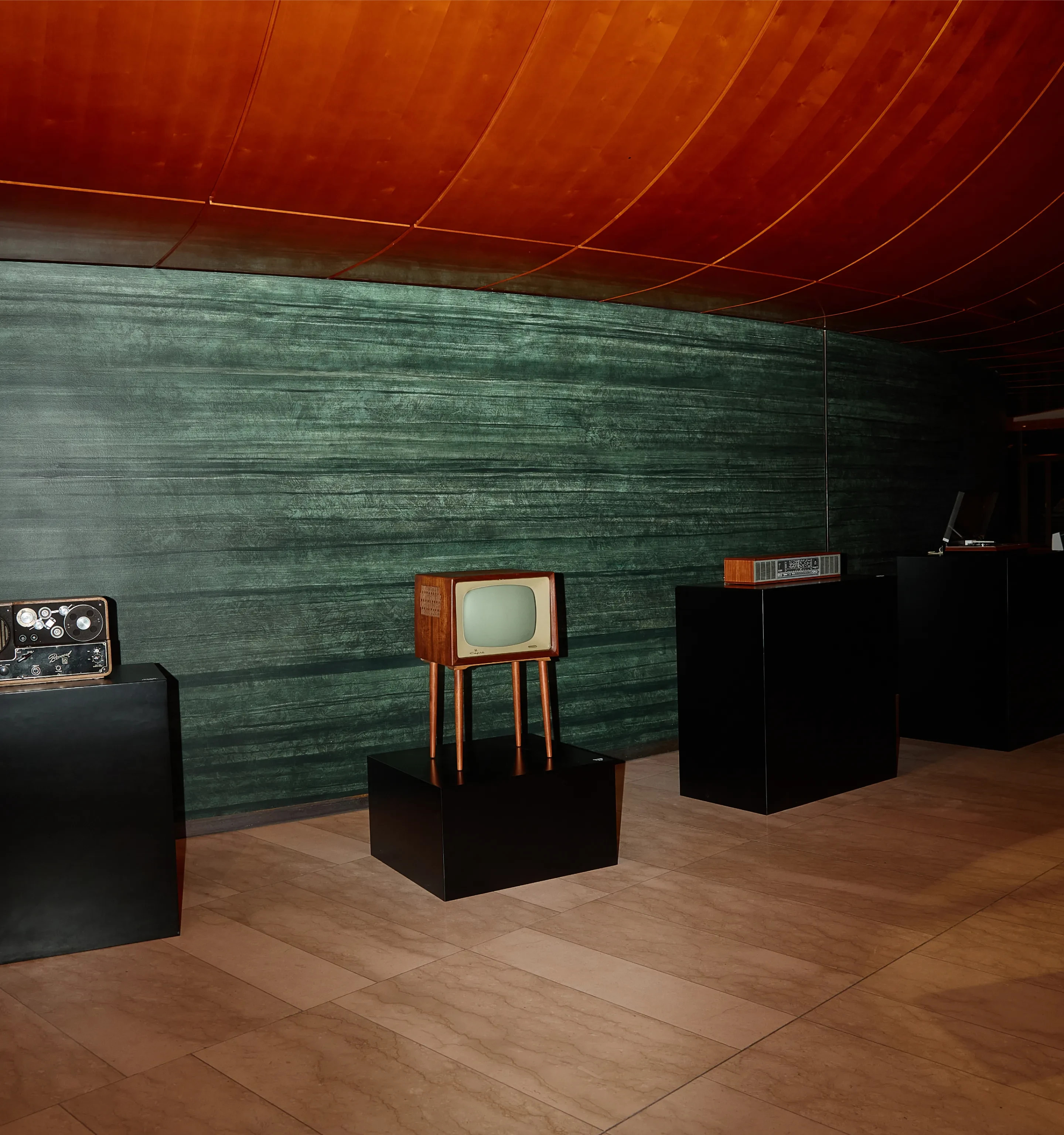 Vintage television and electronics displayed on black pedestals in a room with green walls and warm wooden ceiling.