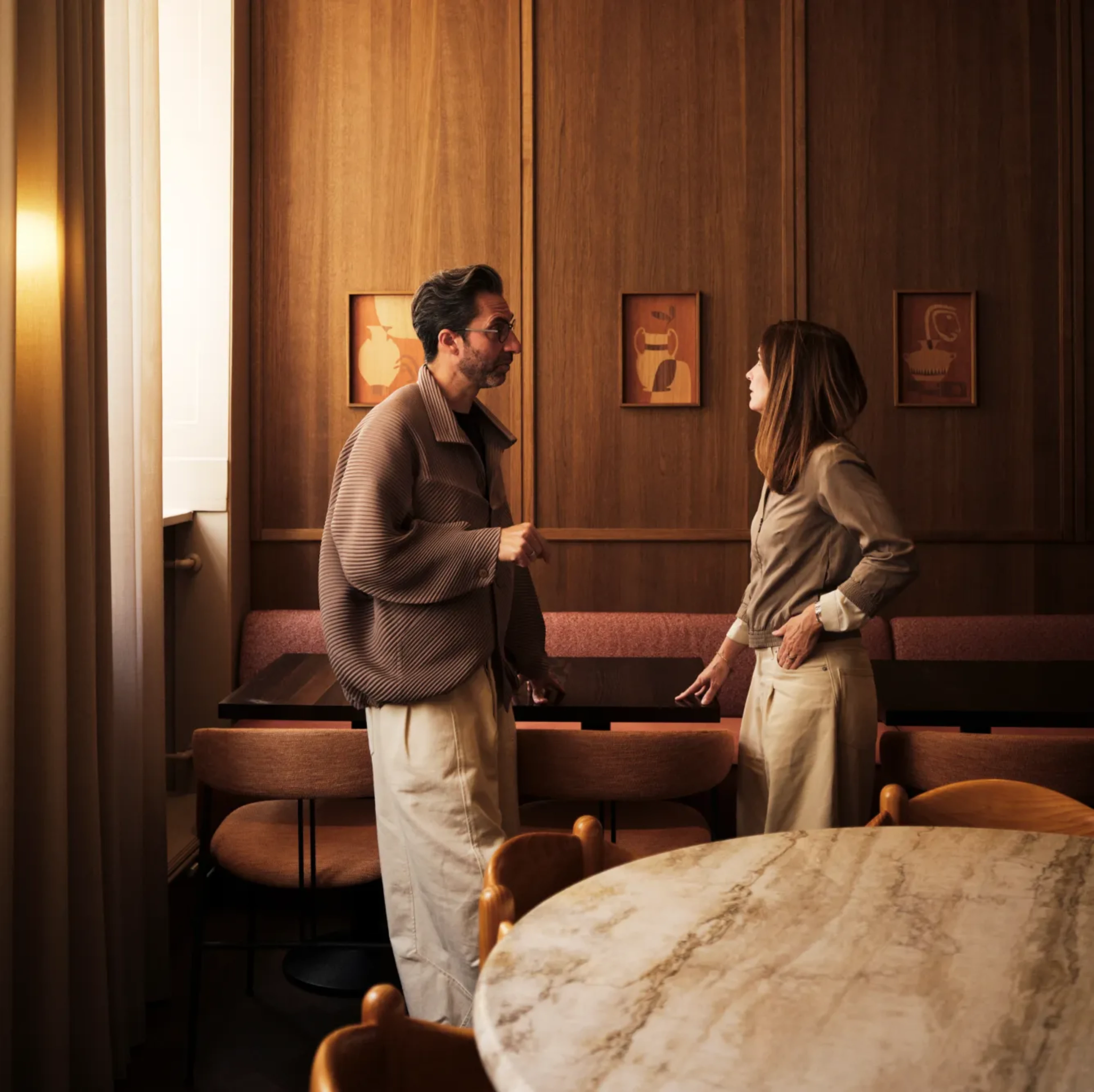Two people stand and converse in a warmly lit dining space featuring dark wood-paneled walls, upholstered seating, and a large round marble table in the foreground.