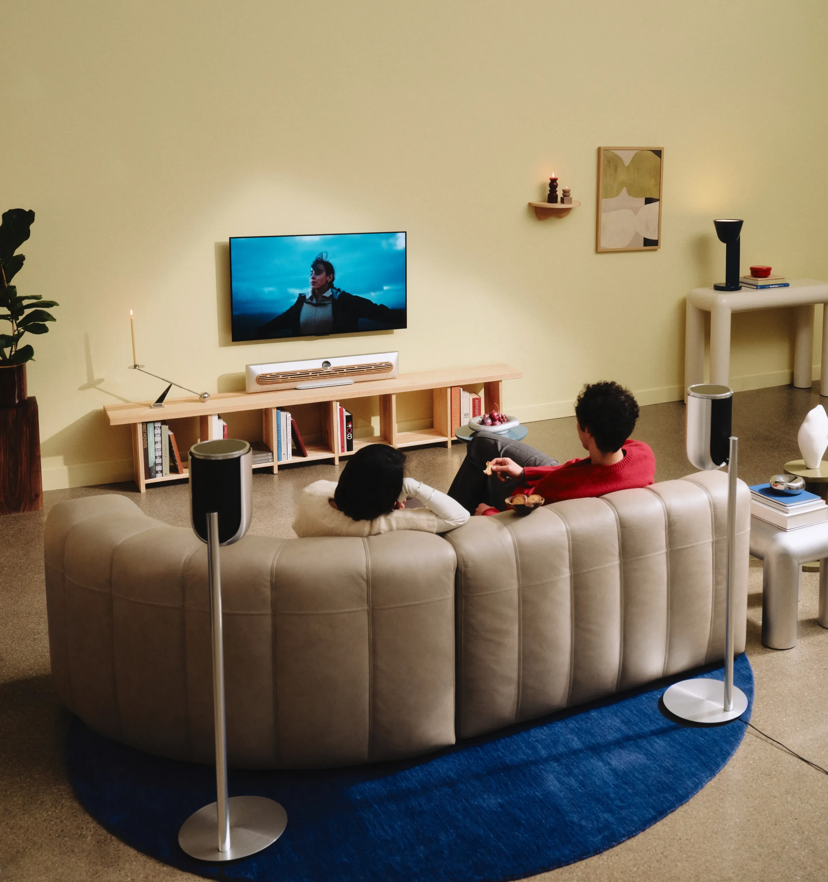 Modern living room with two people relaxing, wall-mounted TV with Beosound Premiere, Beolab 8 surround speakers, and contemporary furniture and artwork.