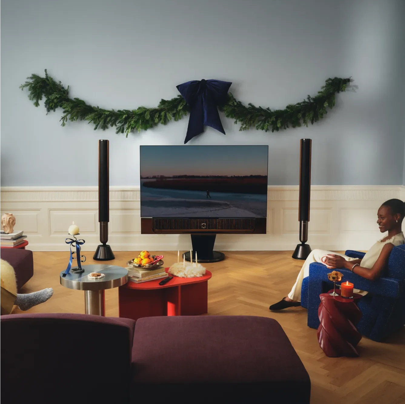 Cozy living room with Beovision Theatre TV displaying sunset beach scene, evergreen garland with blue bow, and person relaxing with a drink.