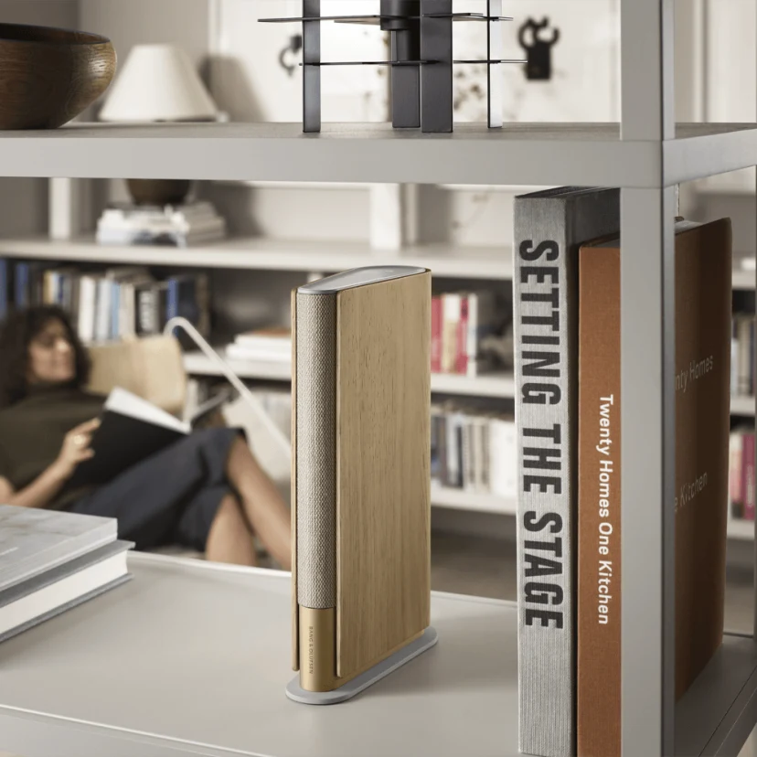 Book-like speaker Beosound Emerge Light Oak placed on a bookshelf, while a woman listens to music and reads in the background.