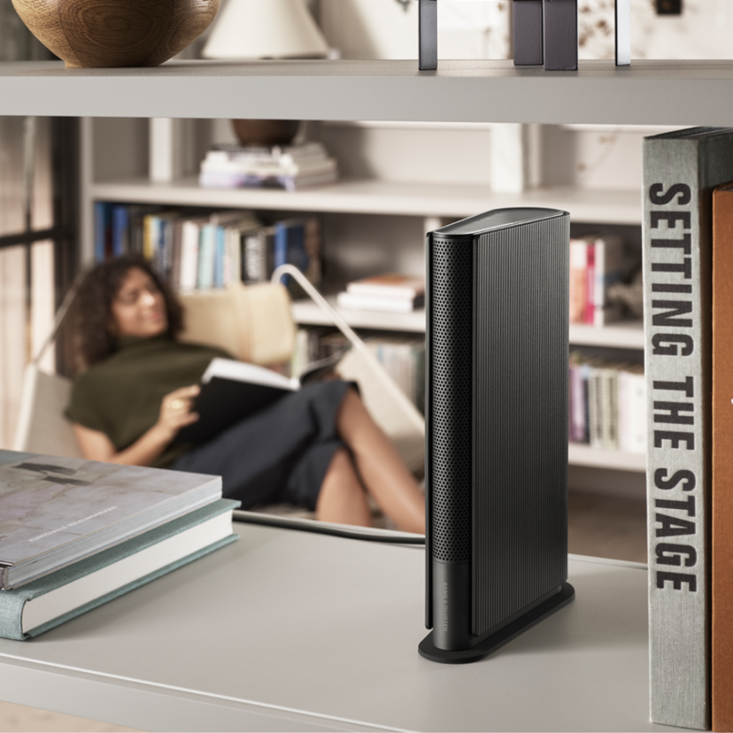 Book-like speaker Beosound Emerge Light Oak placed on a bookshelf, while a woman listens to music and reads in the background.